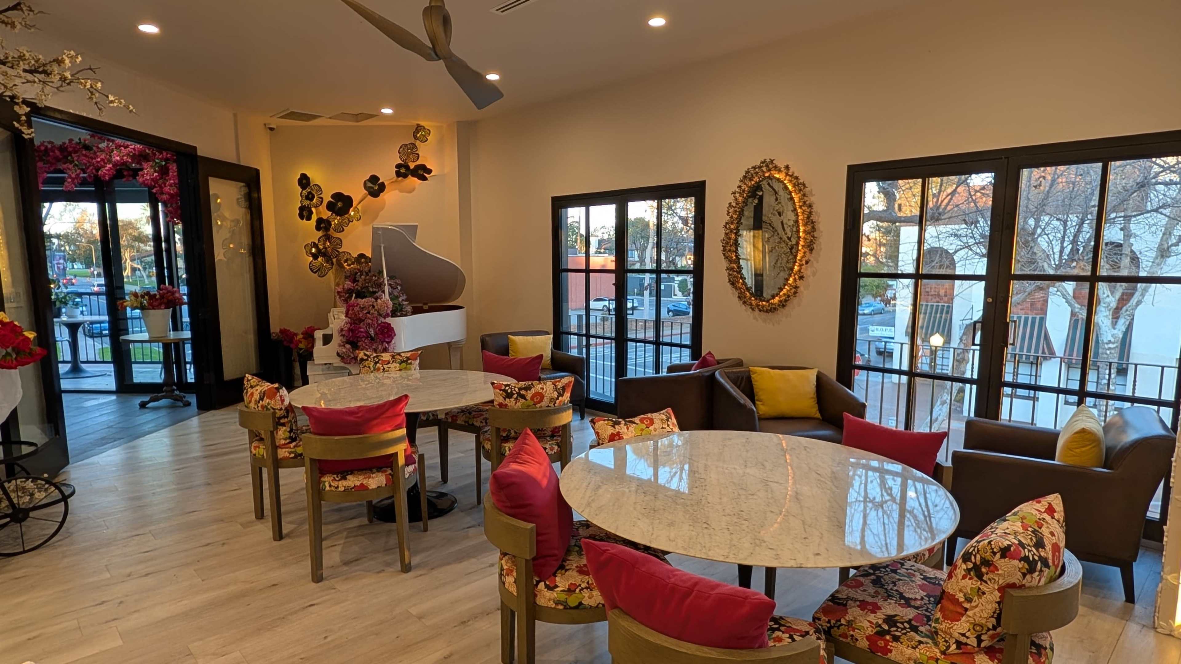 The image shows a modern interior space with several round tables surrounded by colorful chairs, accented by floral decor and large windows.