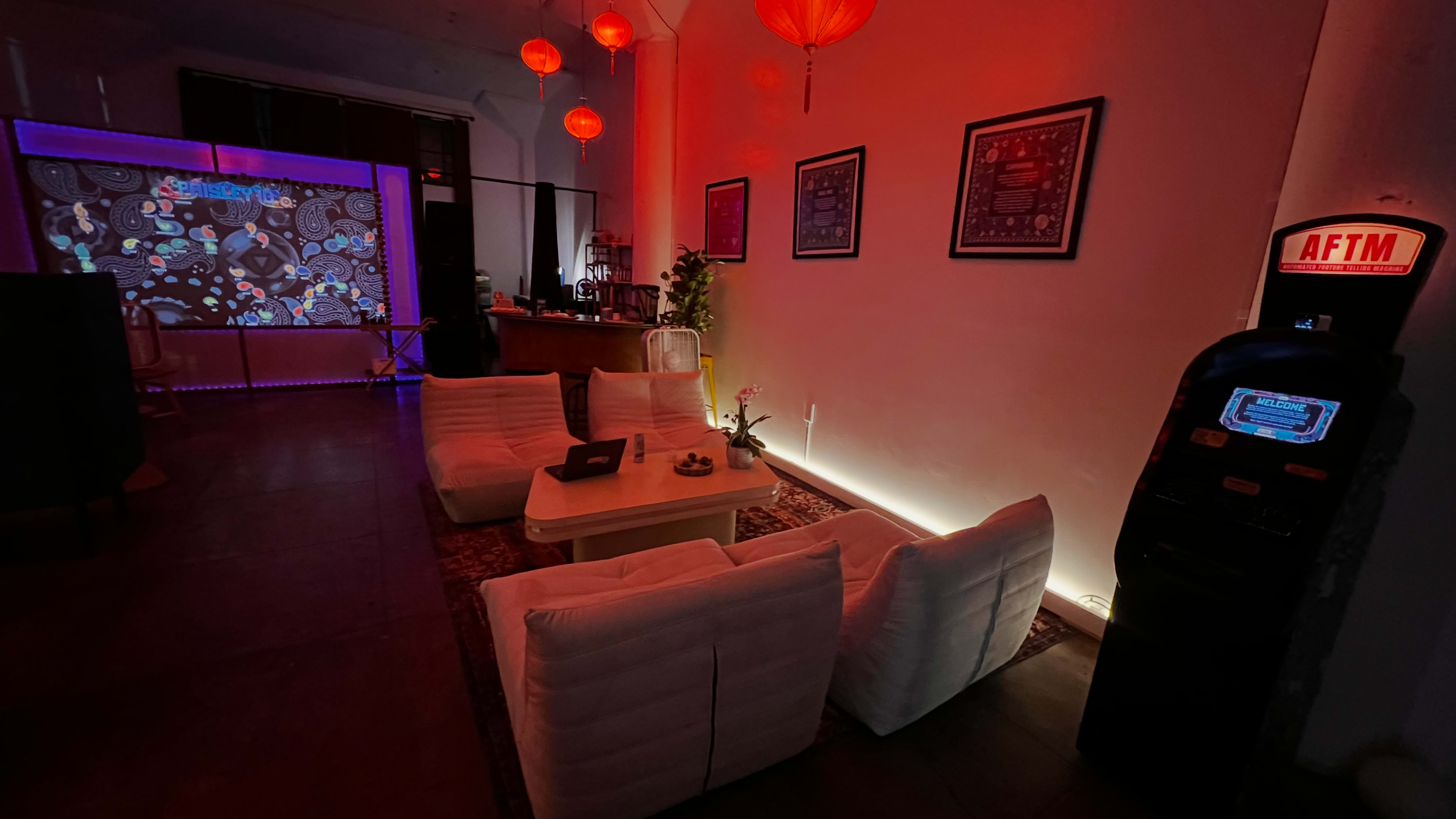 The image shows a dimly lit lounge area with white sofas, a small table in the center, and red lanterns hanging from the ceiling, alongside a large projected screen and a vintage arcade game machine.