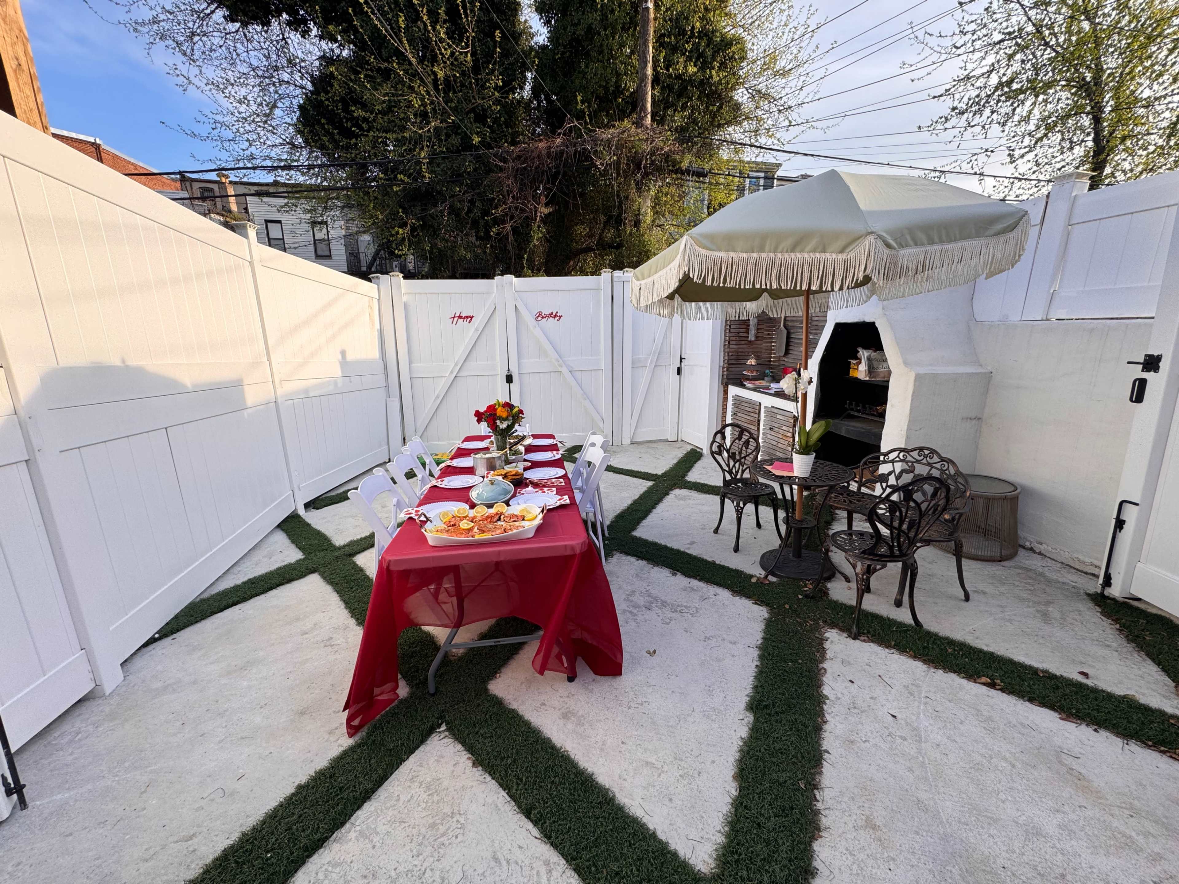 A neatly arranged outdoor seating area features a red tablecloth with a spread of food, surrounded by white fencing and a small covered seating space.