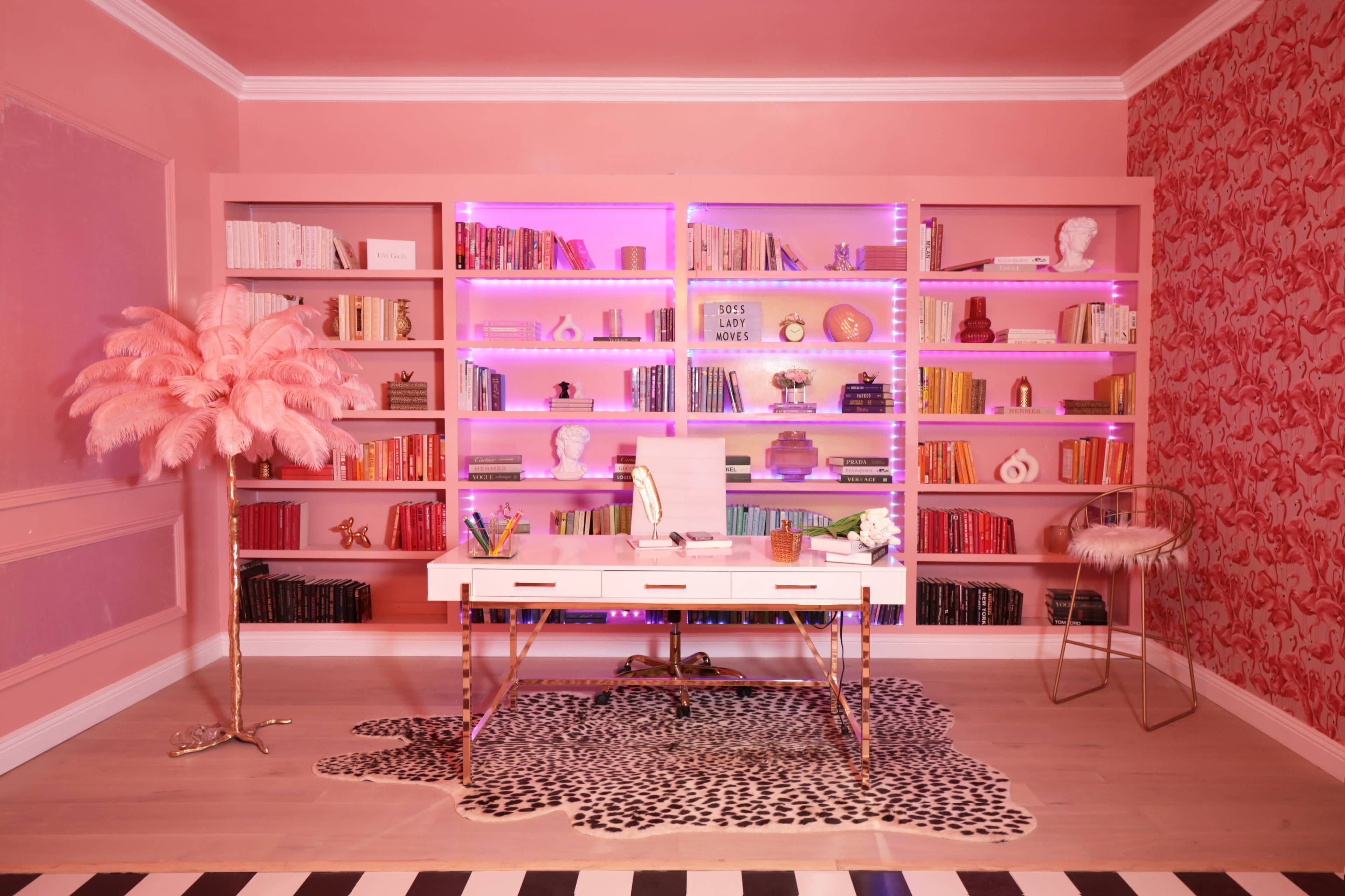 A pink-themed office features a white desk with a mirror, illuminated bookshelves filled with books and decorative items, and a stylish rug on the floor.
