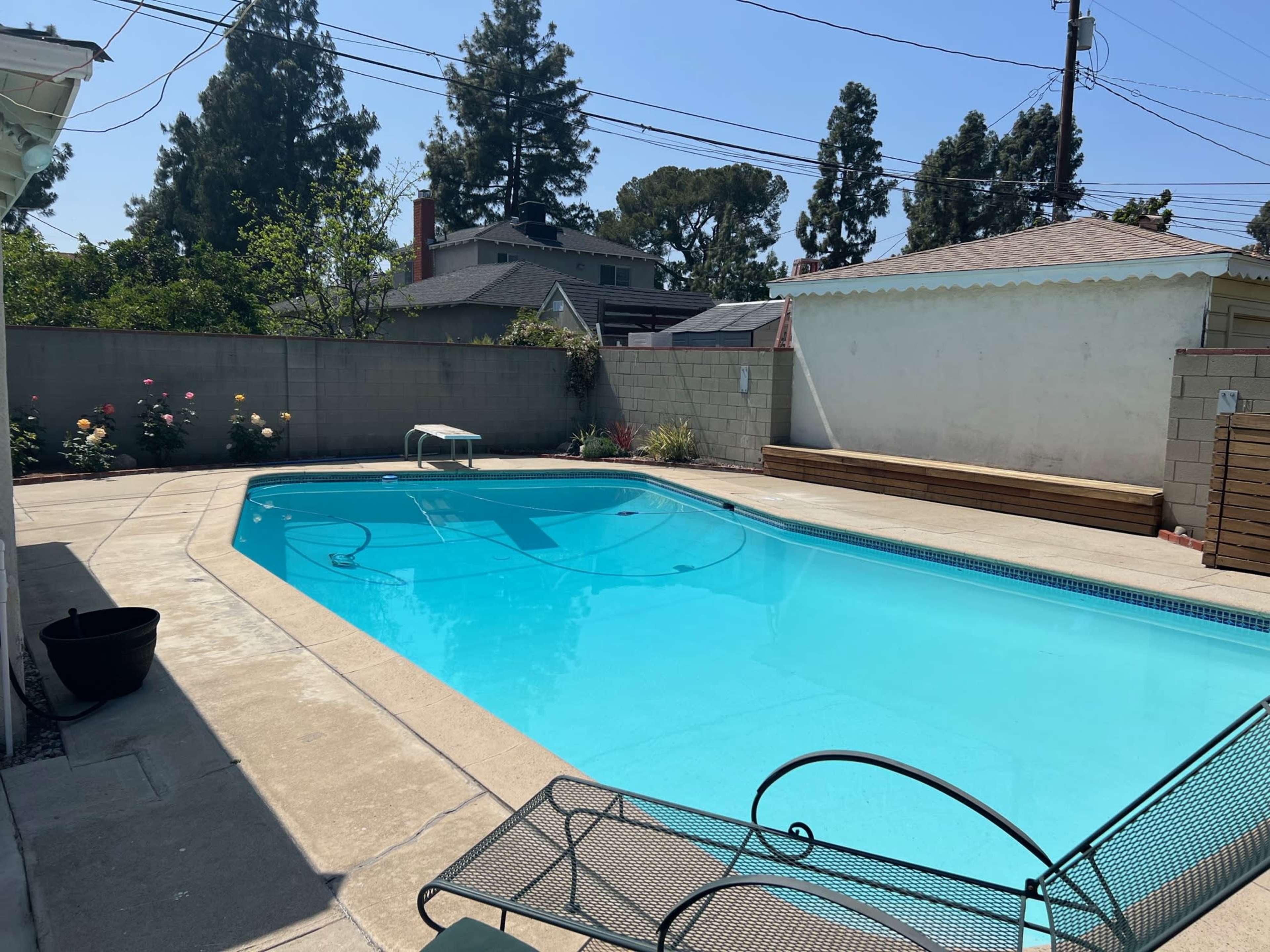 The image shows a clear, rectangular swimming pool surrounded by a concrete deck, with chairs and wooden benches along one side and flowering plants along the perimeter.