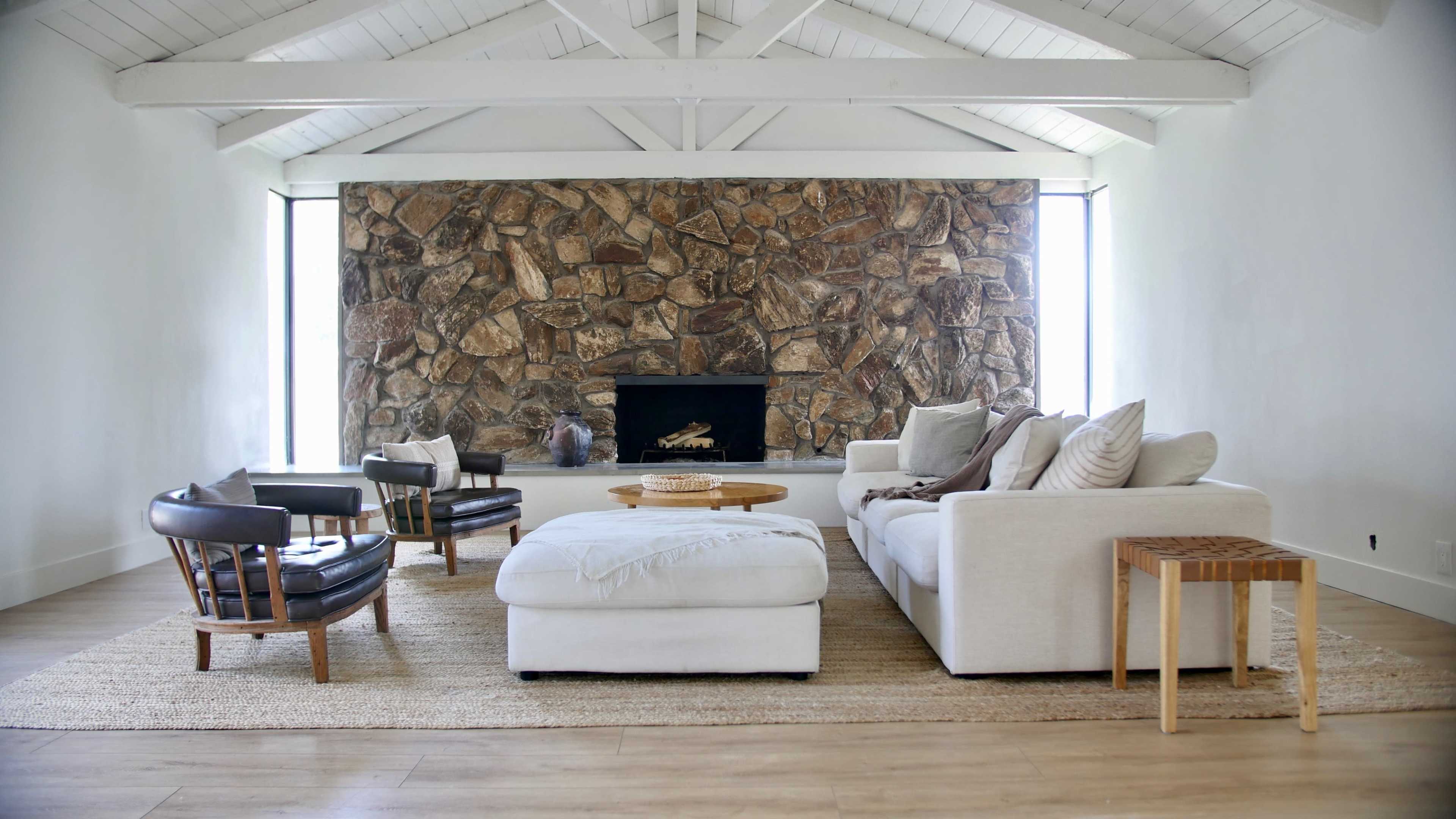 A spacious living room features a large stone fireplace, a white sofa, a round coffee table, and several armchairs arranged around a woven rug.