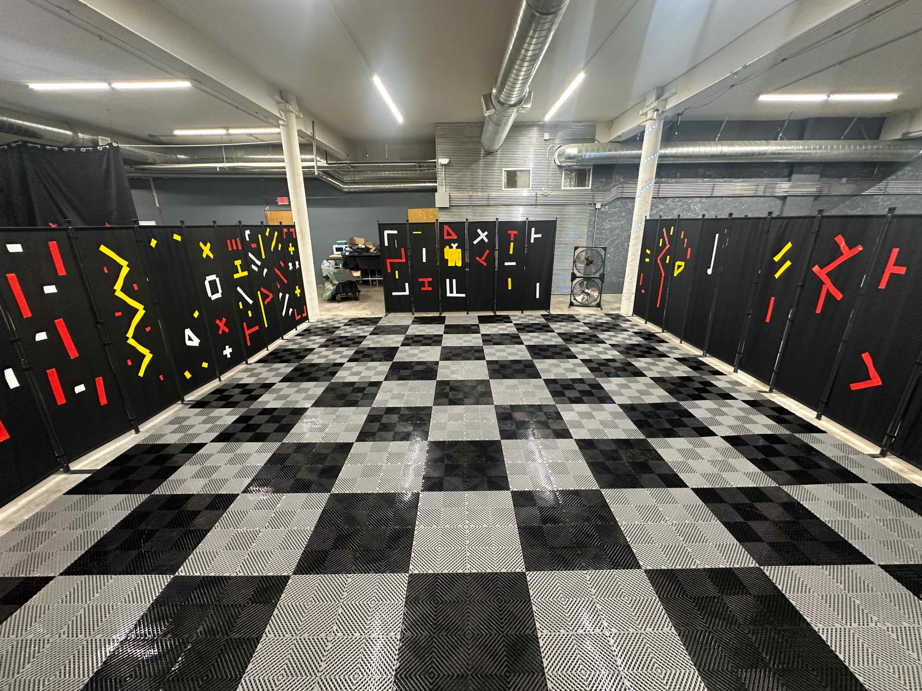 A large room with a checkered black and gray floor, surrounded by black panels adorned with red, yellow, and white geometric shapes.