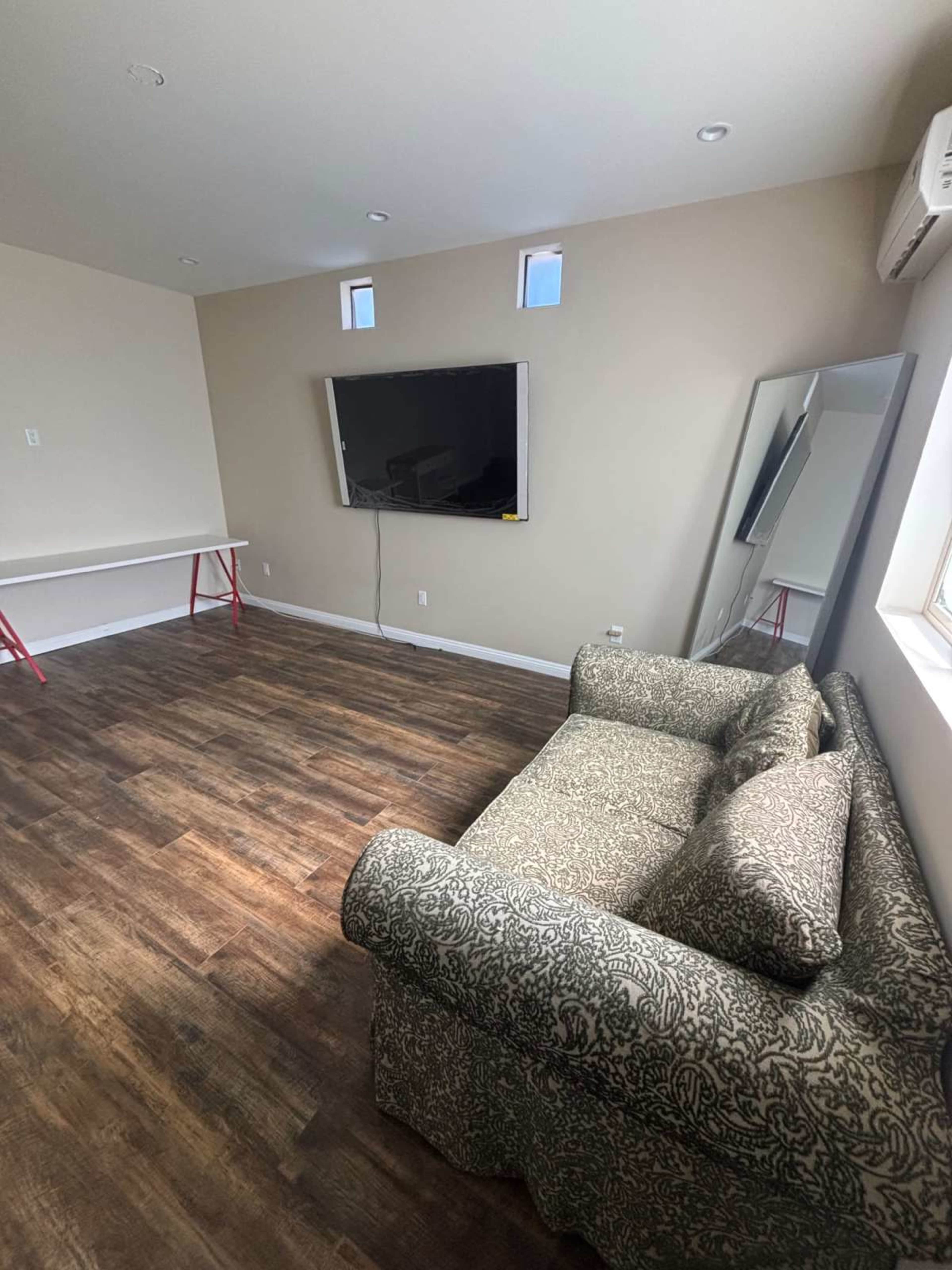 The room features a couch, a wall-mounted television, a mirror, and a table with red legs on a wooden floor.