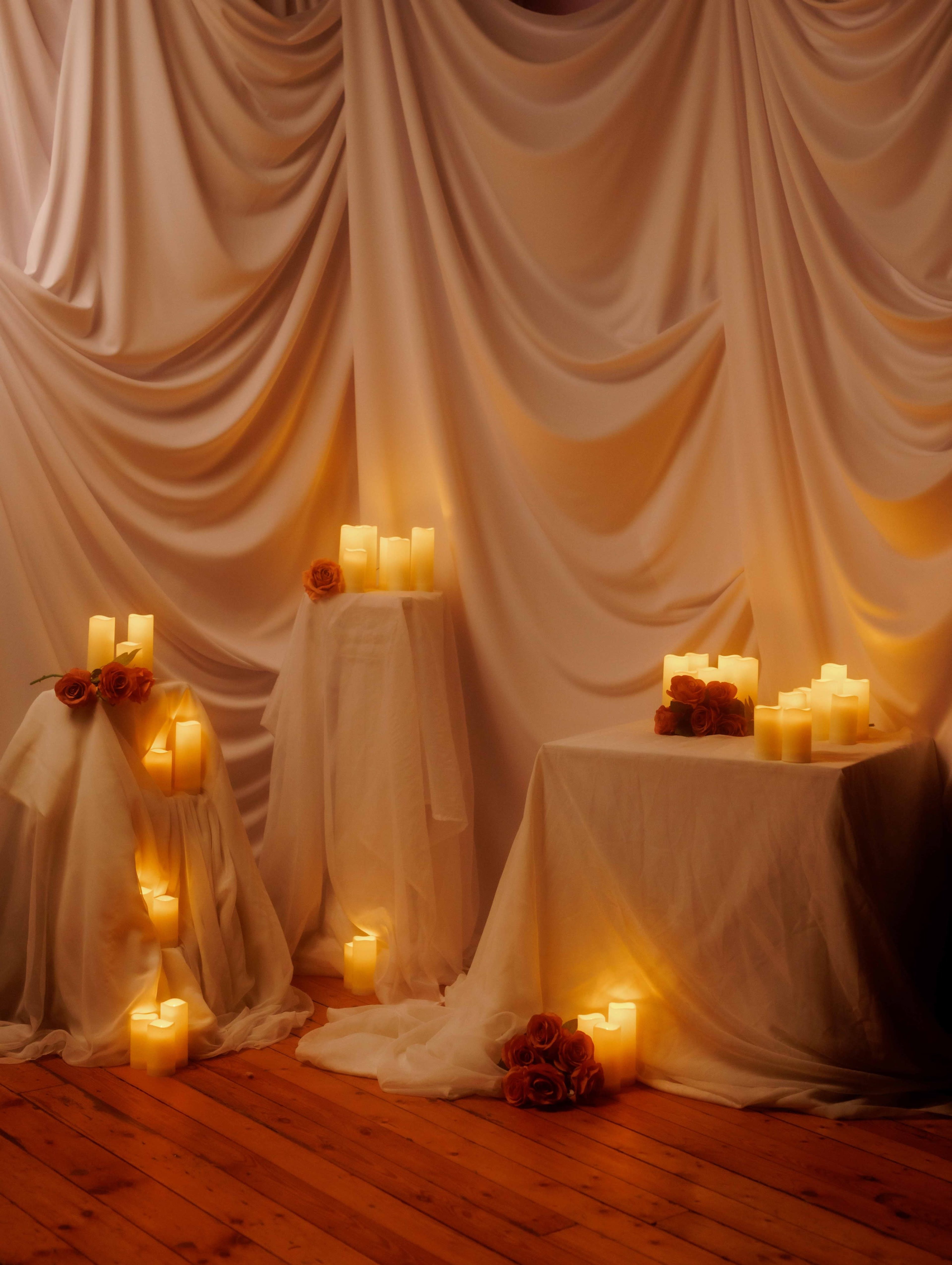The scene features three white draped tables adorned with numerous candle arrangements and roses against a soft, textured backdrop.