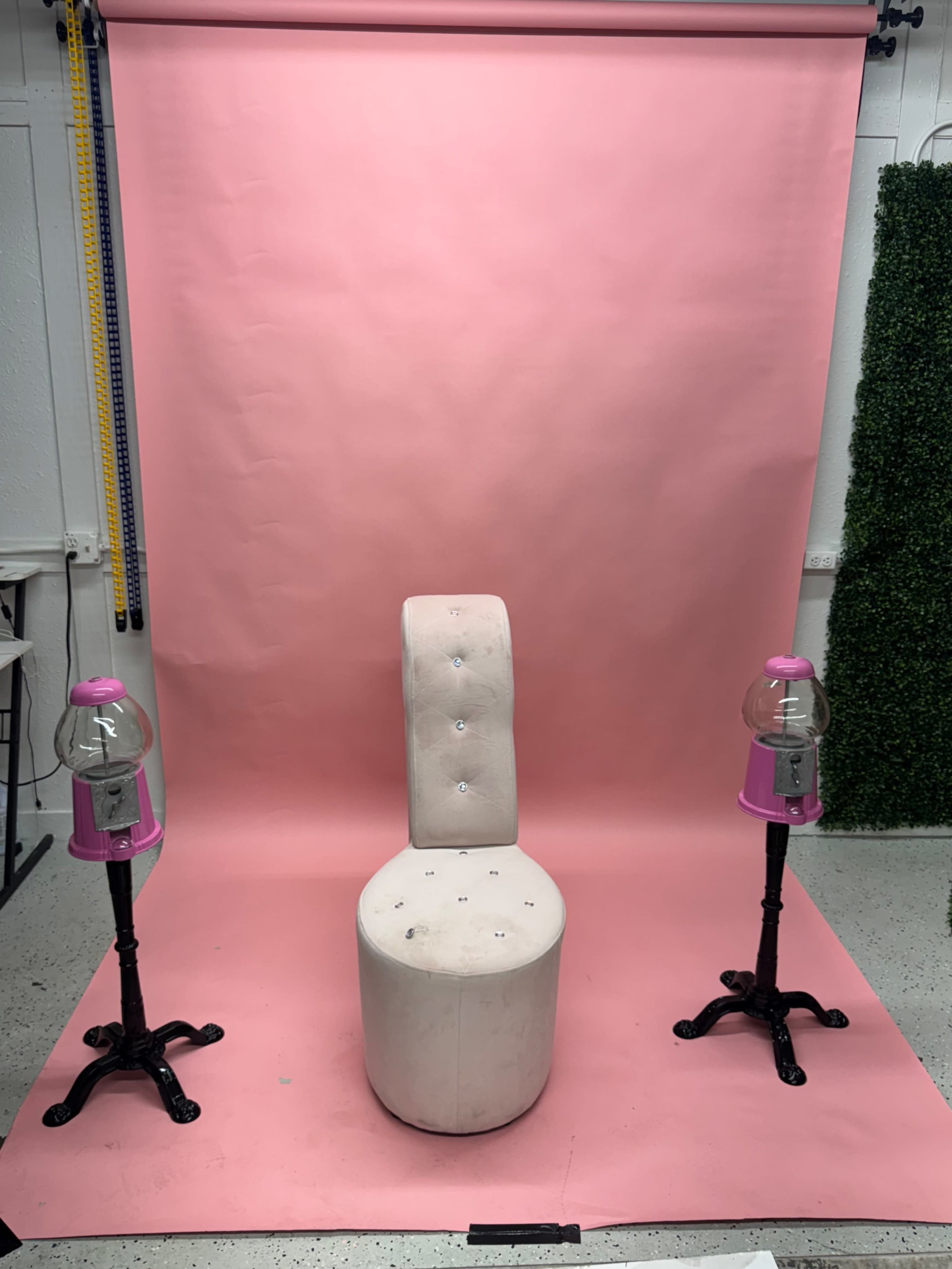 The image shows a light pink backdrop with a white, embellished chair in the center, flanked by two pink gumball machines on black stands.