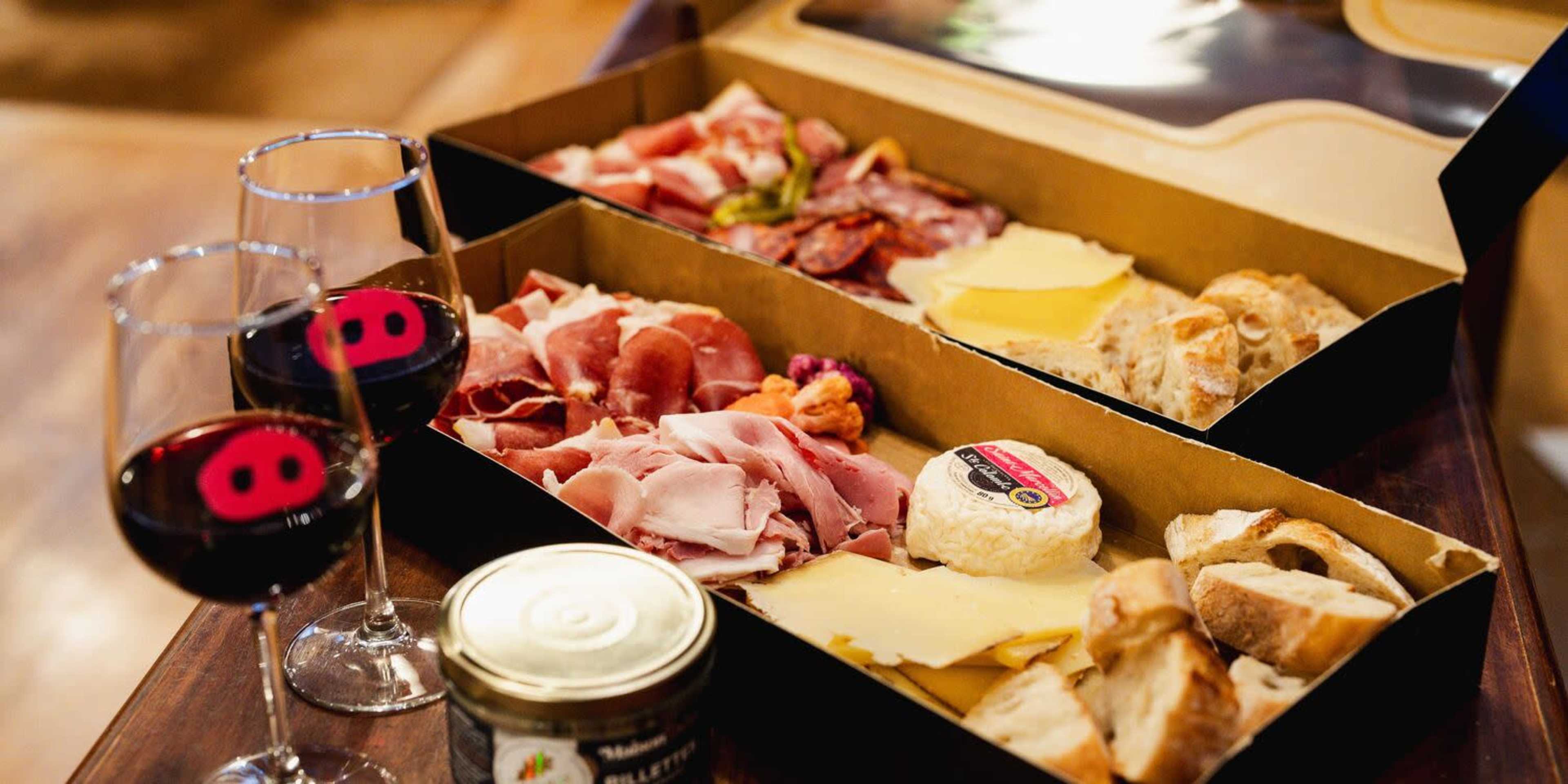 The image shows a wooden table with a charcuterie board featuring various meats, cheeses, breads, and wine glasses.