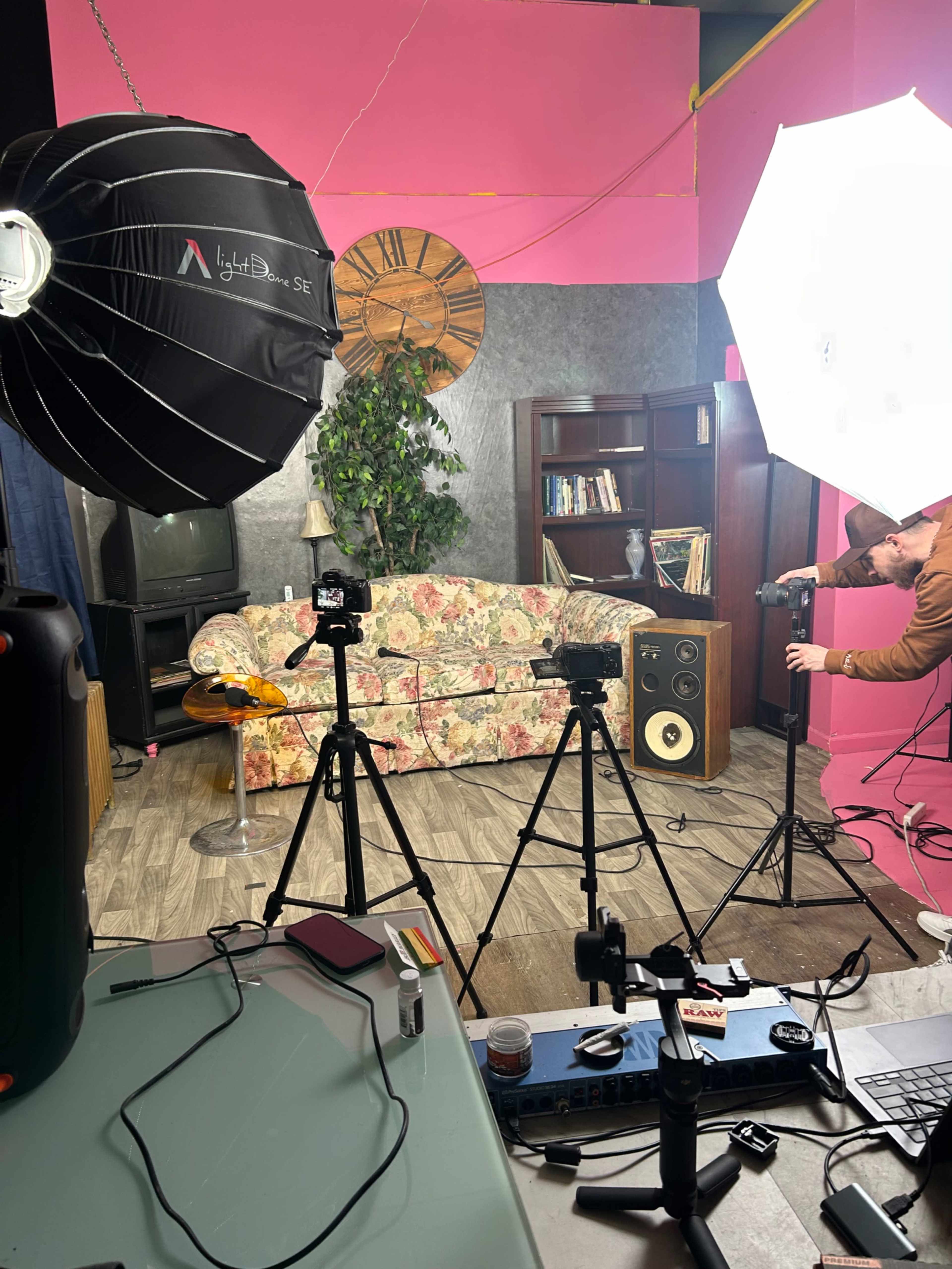 The image shows a studio setup with a floral couch, a large softbox light, a few tripods with cameras, a bookshelf, and a potted plant.
