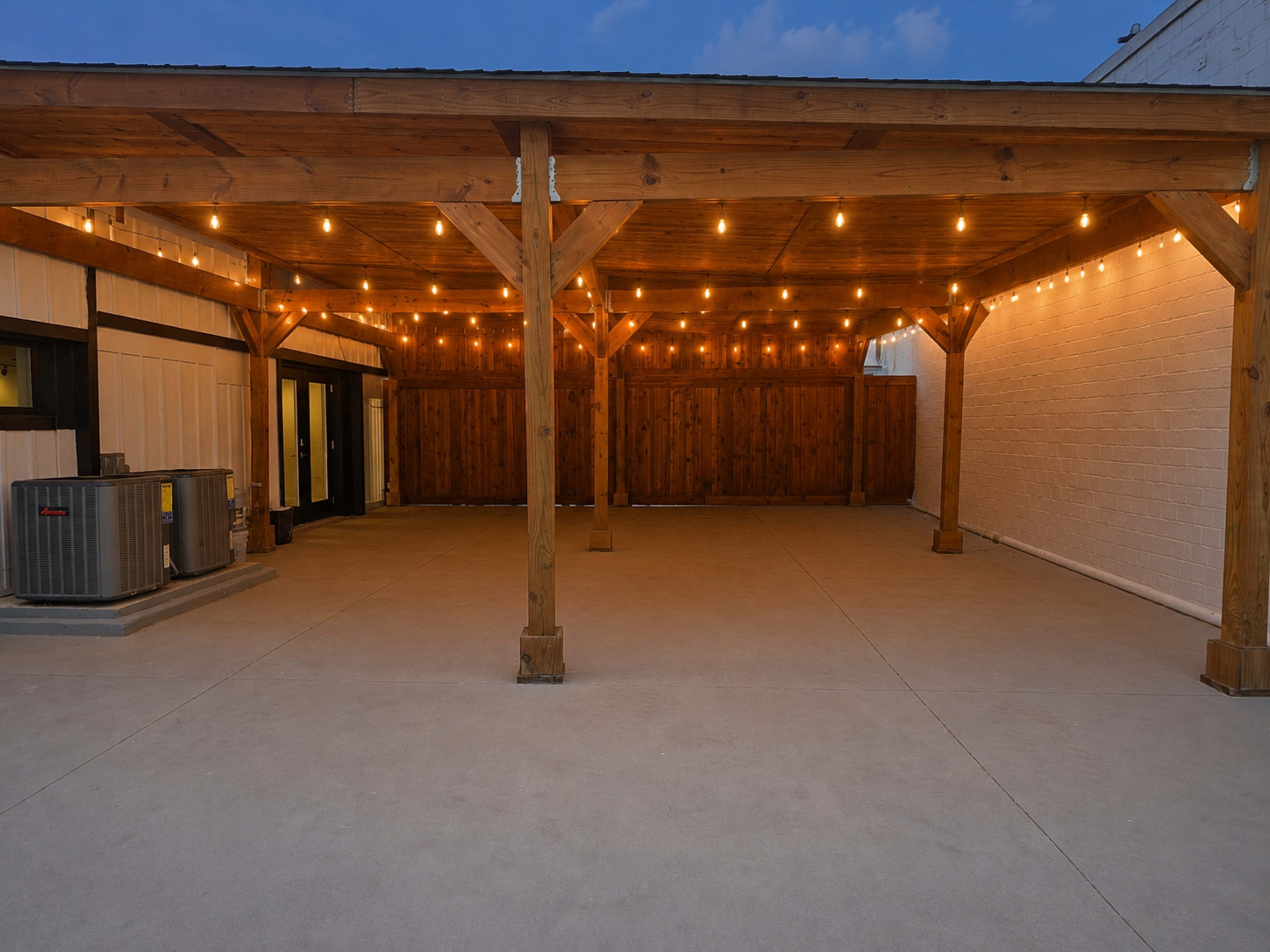 The image shows a spacious outdoor area with wooden beams, string lights, and a concrete floor, enclosed by a wooden fence.