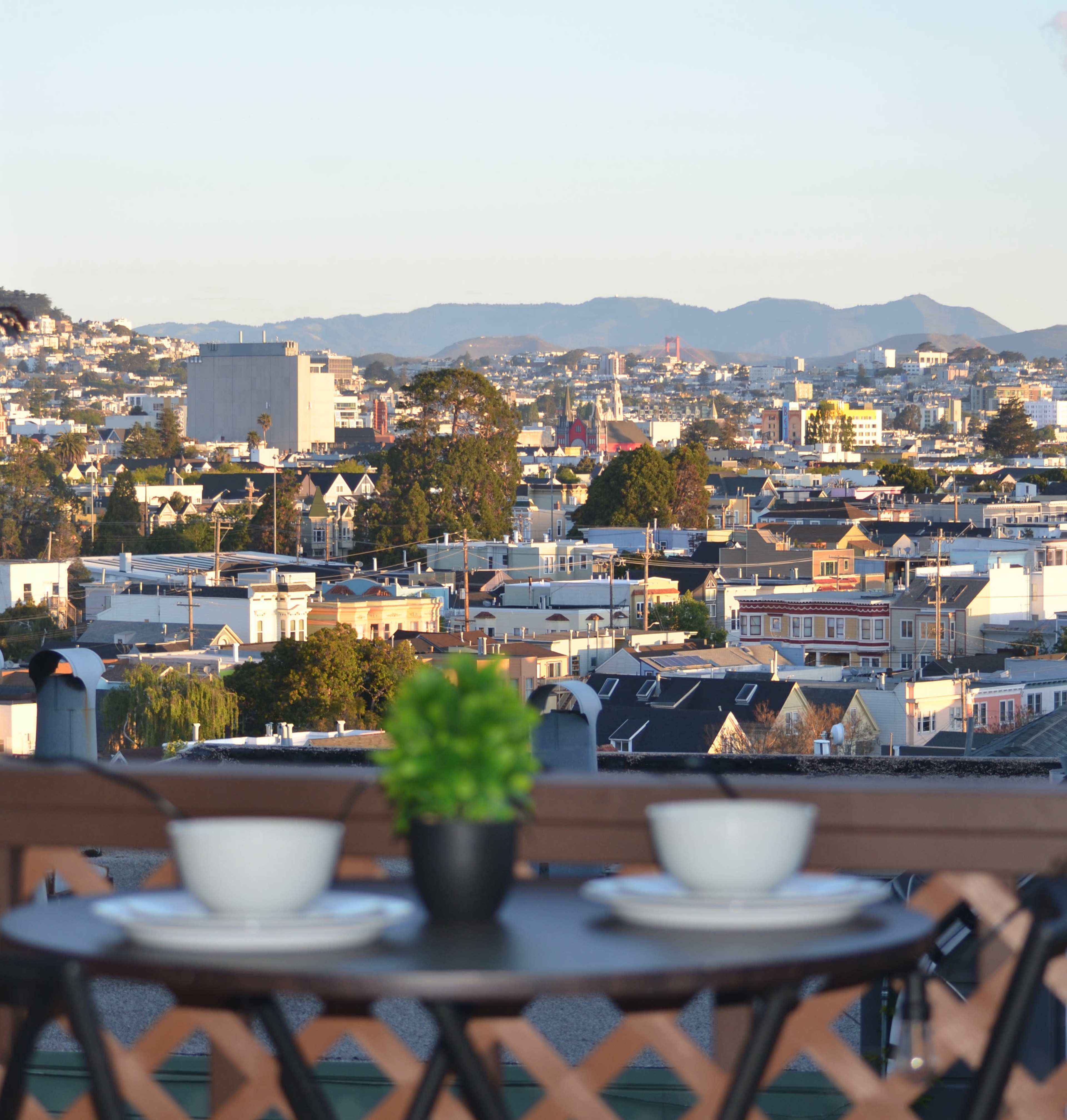 A table set for two with a small plant overlooks a cityscape of rooftops and distant mountains.