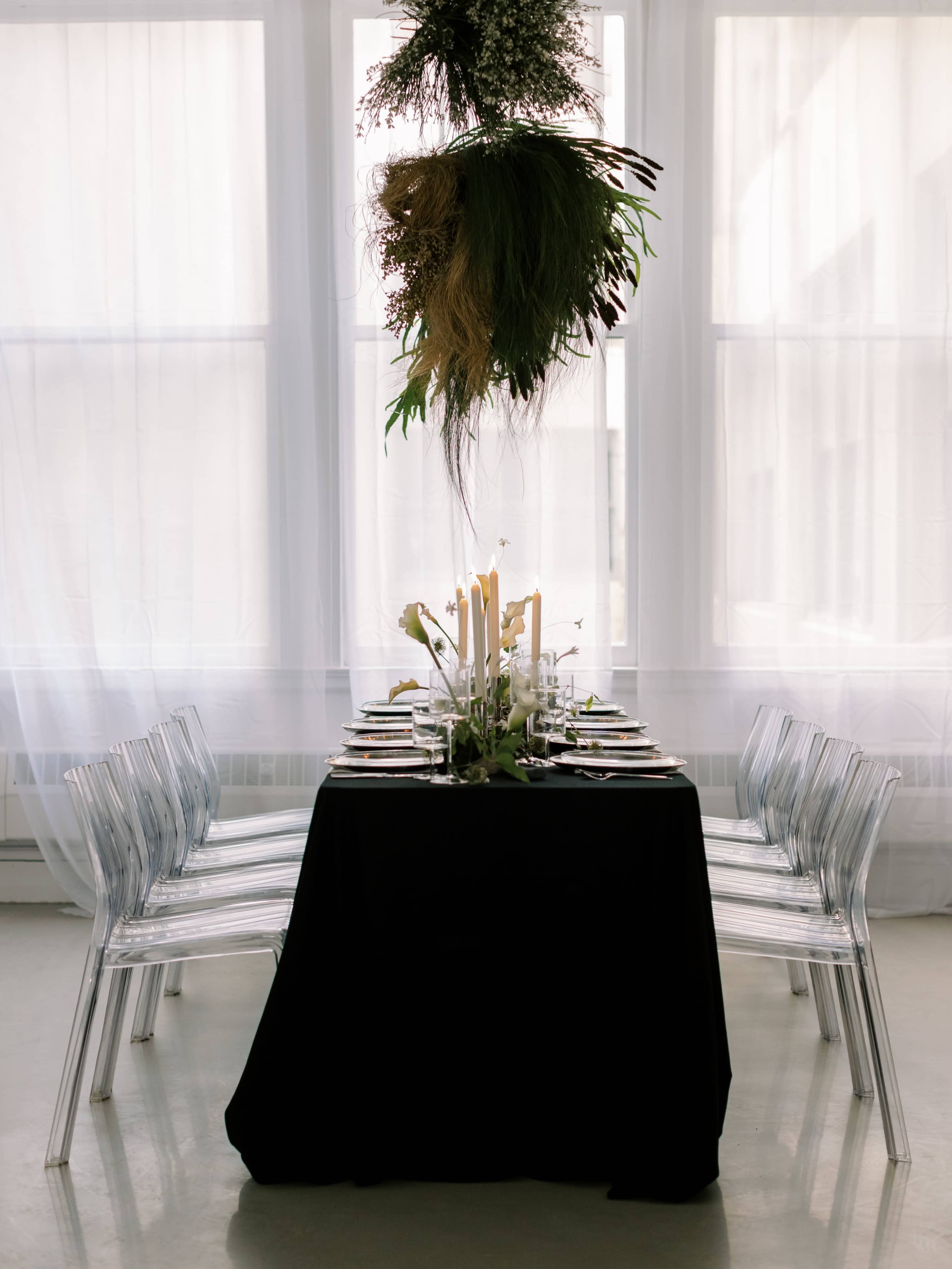 A dining table is set with elegant tableware and surrounded by transparent chairs, with a floral arrangement hanging above in a well-lit room.