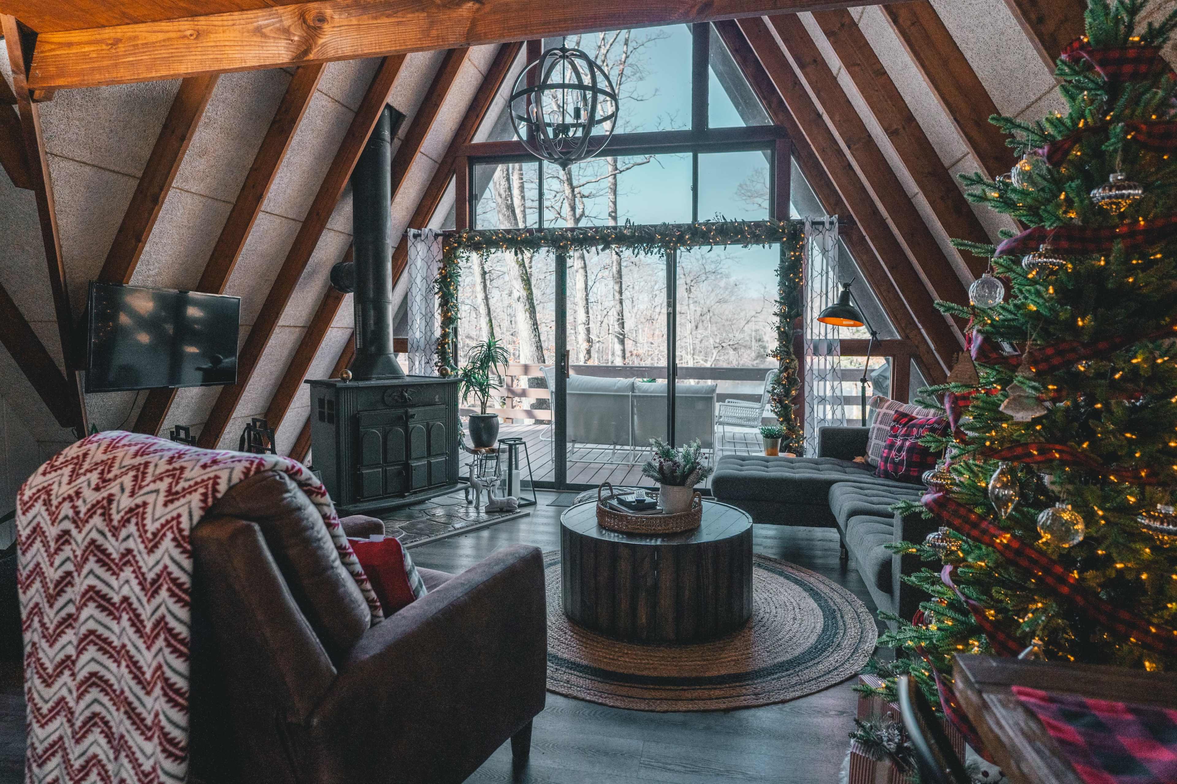 The image depicts a cozy living room with an A-frame structure, featuring a fireplace, a large window with a winter view, and a decorated Christmas tree.