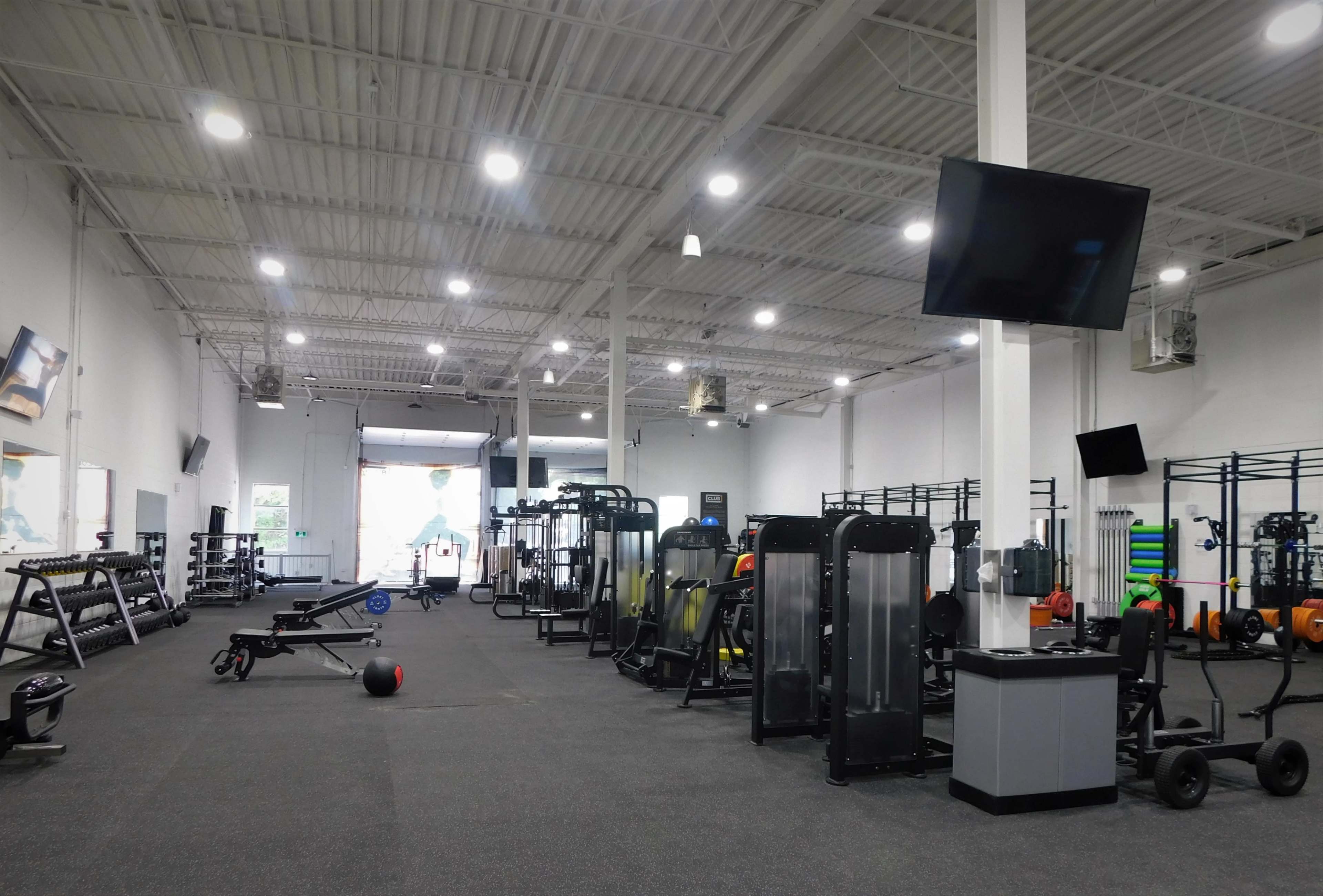 A spacious gym interior with various exercise machines and equipment arranged neatly along the walls, and large windows allowing natural light to enter.