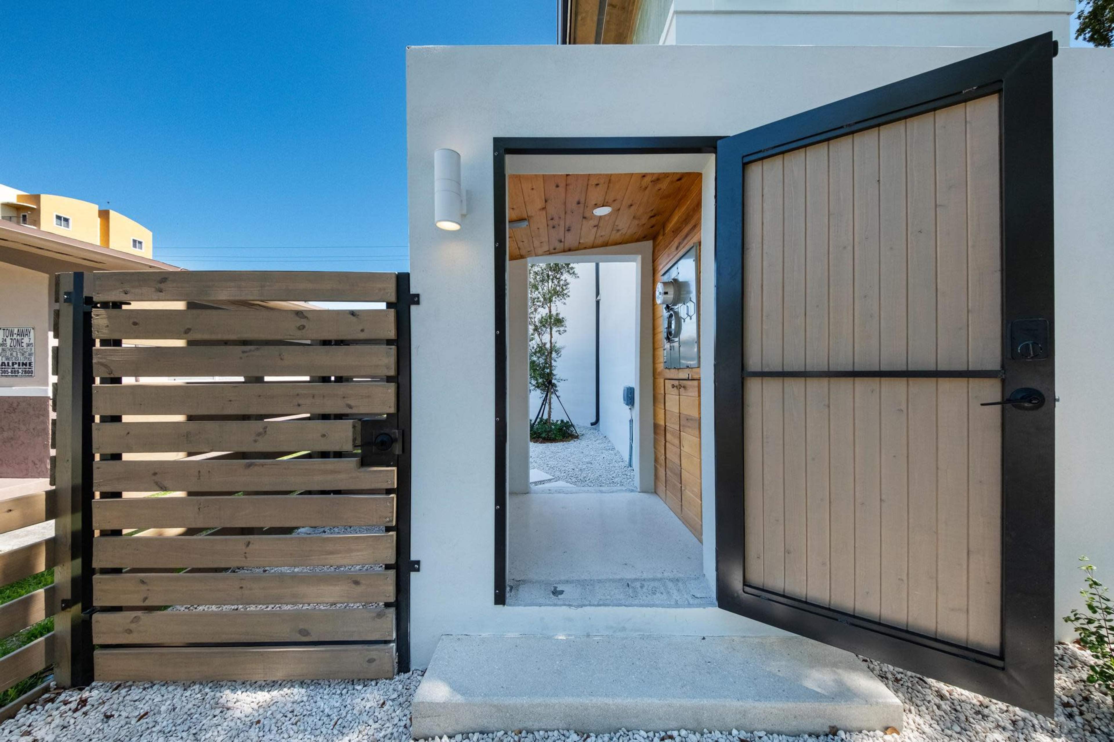 The image shows a stylish entrance with a wooden gate and a door that opens into a well-lit entryway surrounded by white gravel and greenery.