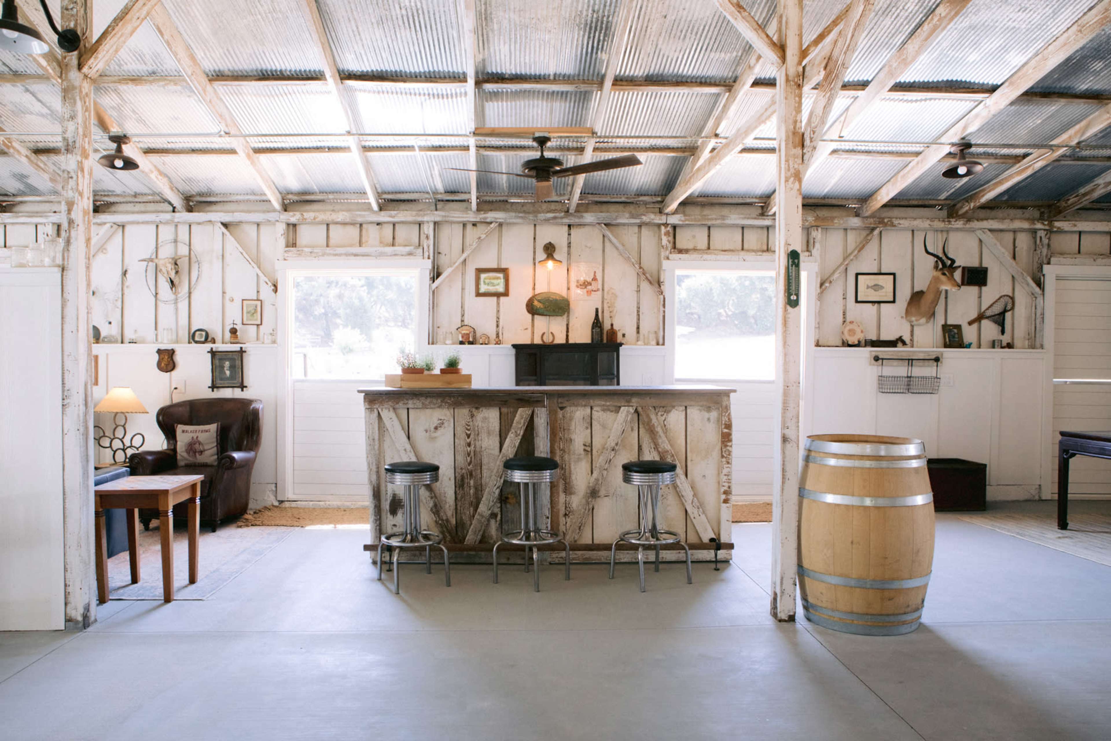 A rustic interior featuring a wooden bar, three metal stools, a barrel, and walls adorned with various decor items.