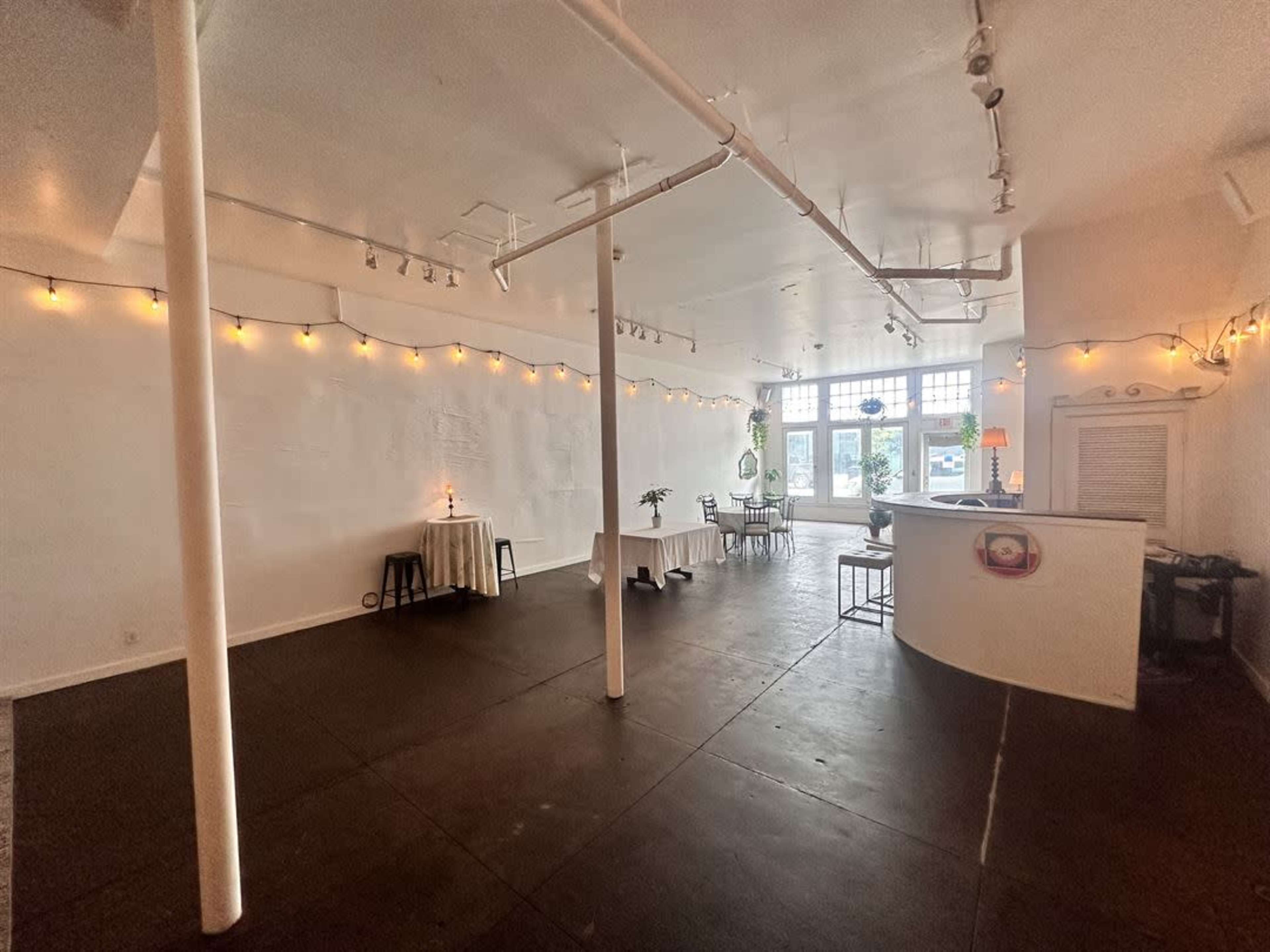 An empty, well-lit interior space with white walls, a dark floor, and a few tables near the windows.