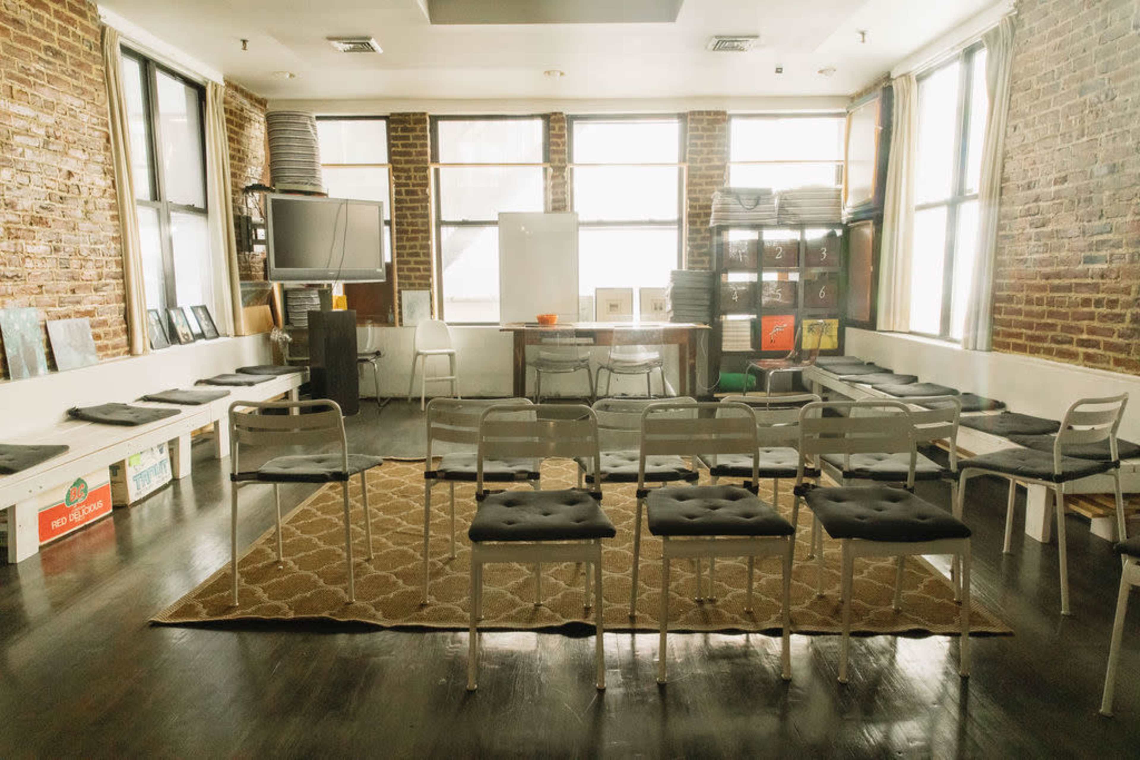 A spacious room with exposed brick walls, featuring a central area of chairs arranged in rows, a table, and various objects along the walls.