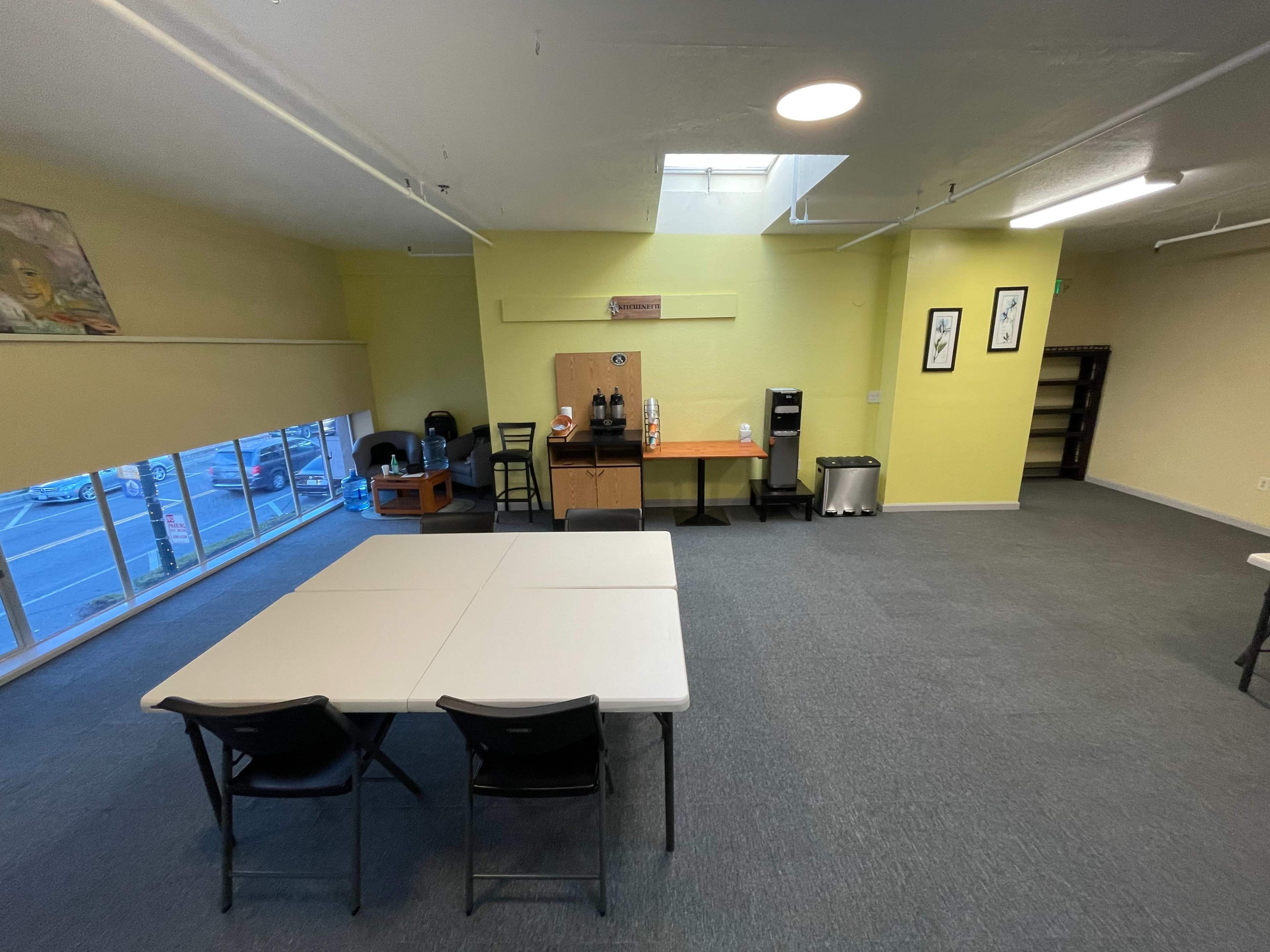 A spacious, brightly lit room featuring a central table with chairs, a coffee station, and large windows overlooking a parking area.