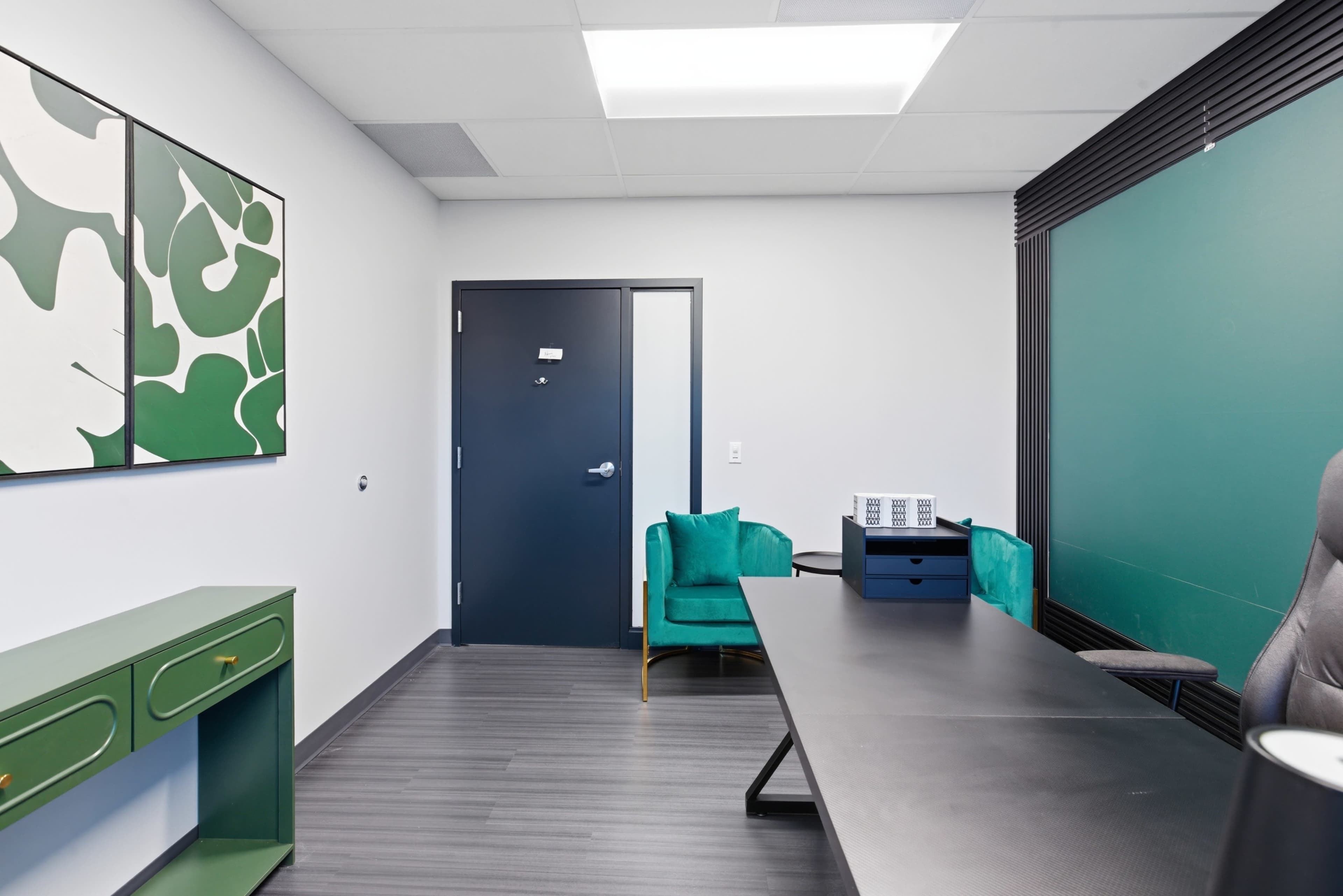 The image shows a modern office space featuring a dark door, a large black table, two green chairs, and a green accent wall with artwork.