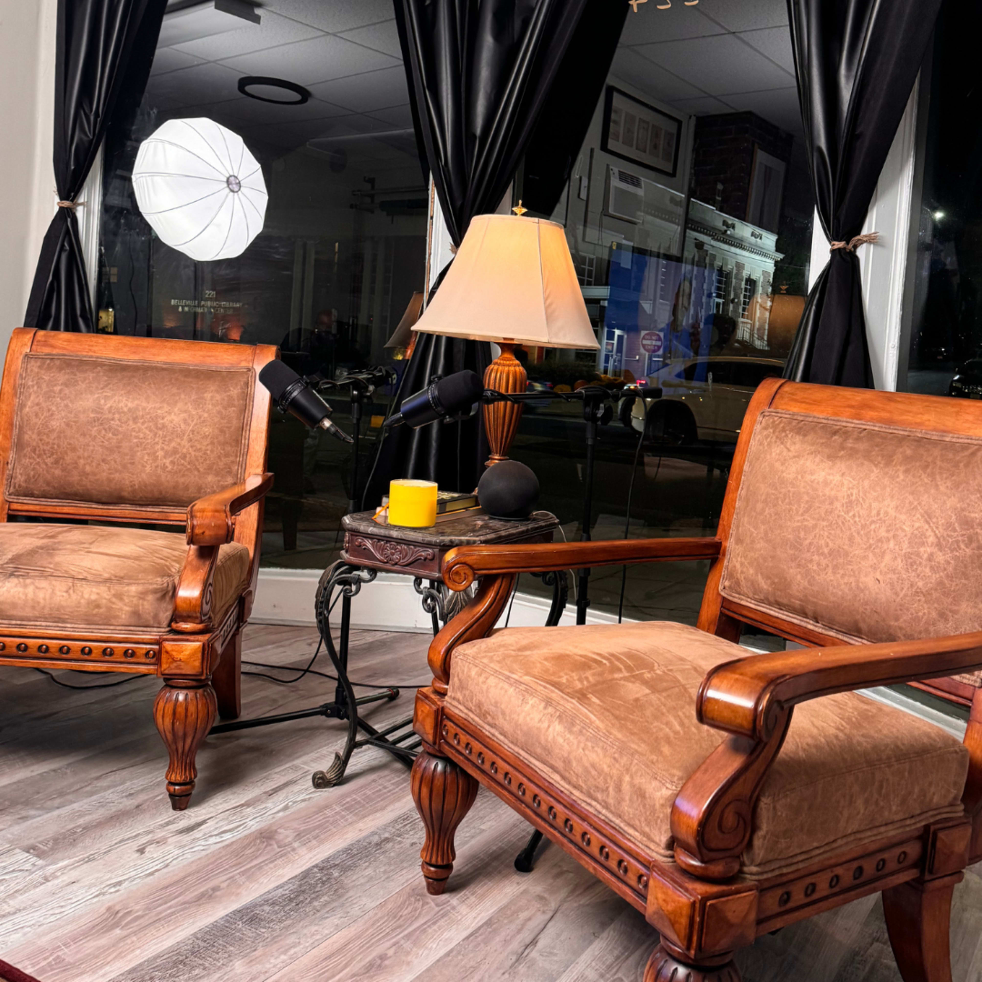 The scene features two wooden chairs with leather upholstery, a lamp on a small round table, and a large window showing an outdoor view in a well-lit room.