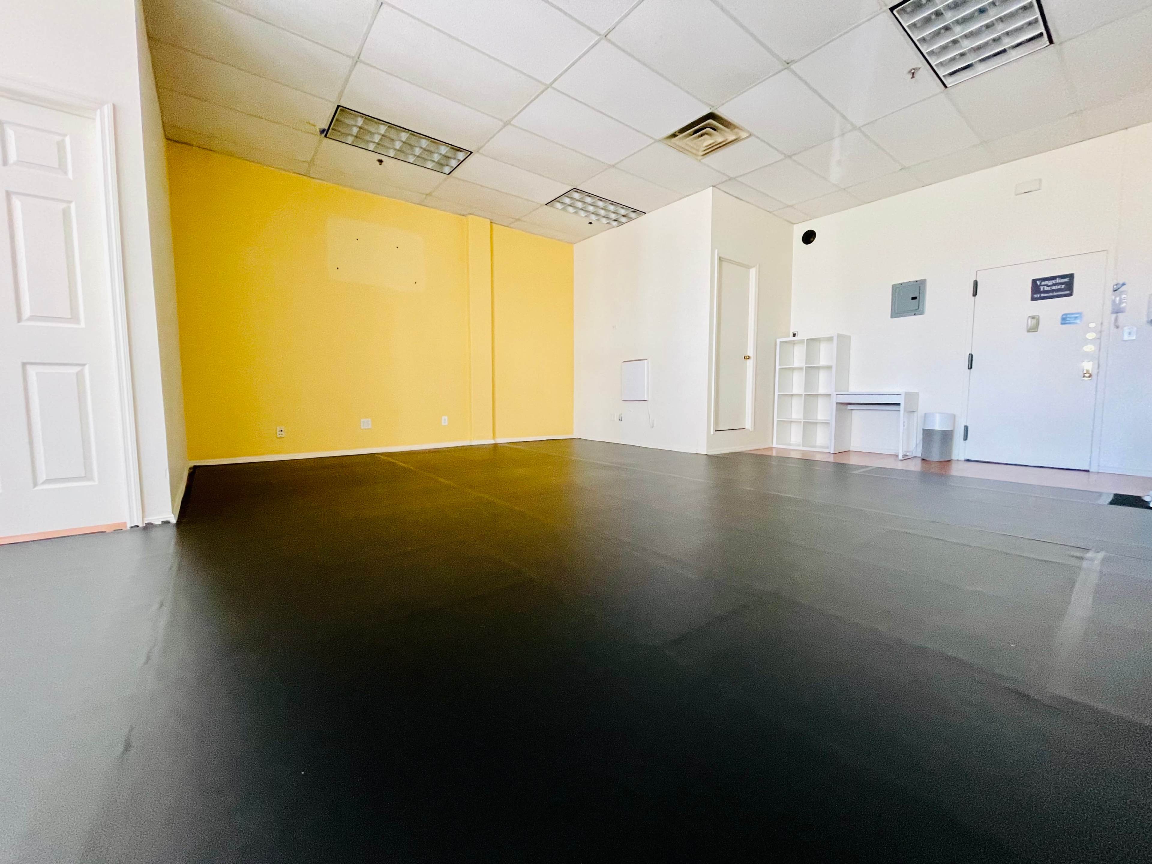 An empty room with a black vinyl floor and yellow walls, featuring a white door and a small shelving unit.