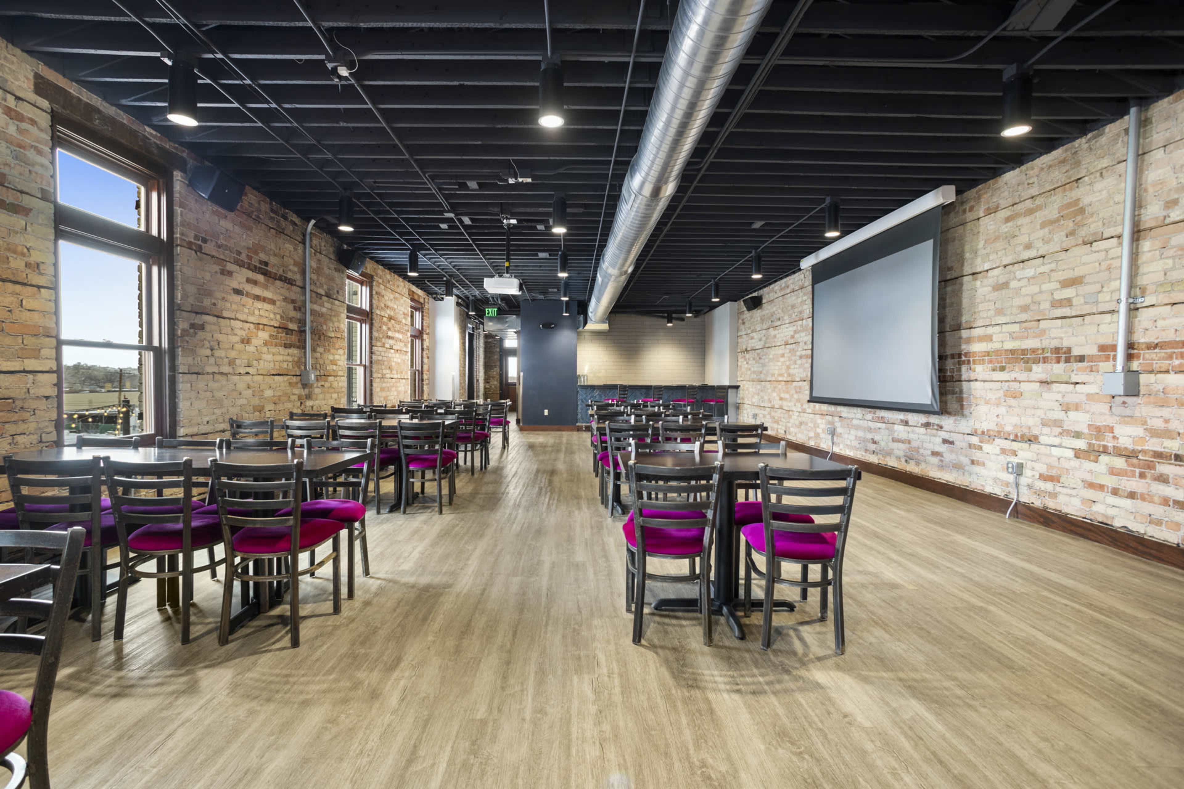 The image shows a large, open dining area with rows of black chairs and pink cushions arranged around wooden tables, brick walls, and a large screen at one end.