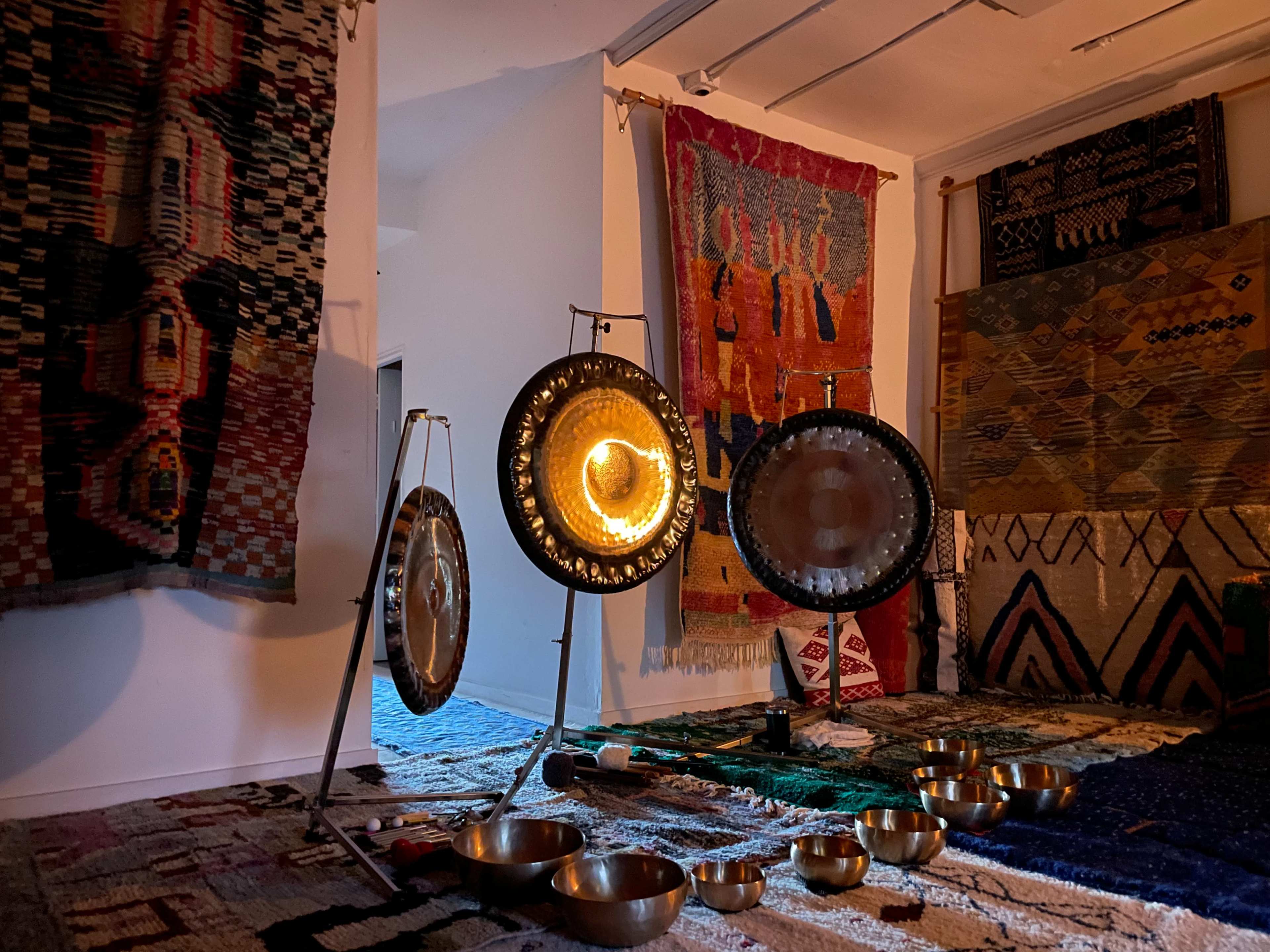 The image shows a room adorned with various textiles on the walls, featuring metallic bowls and gongs set up in a dimly lit space.