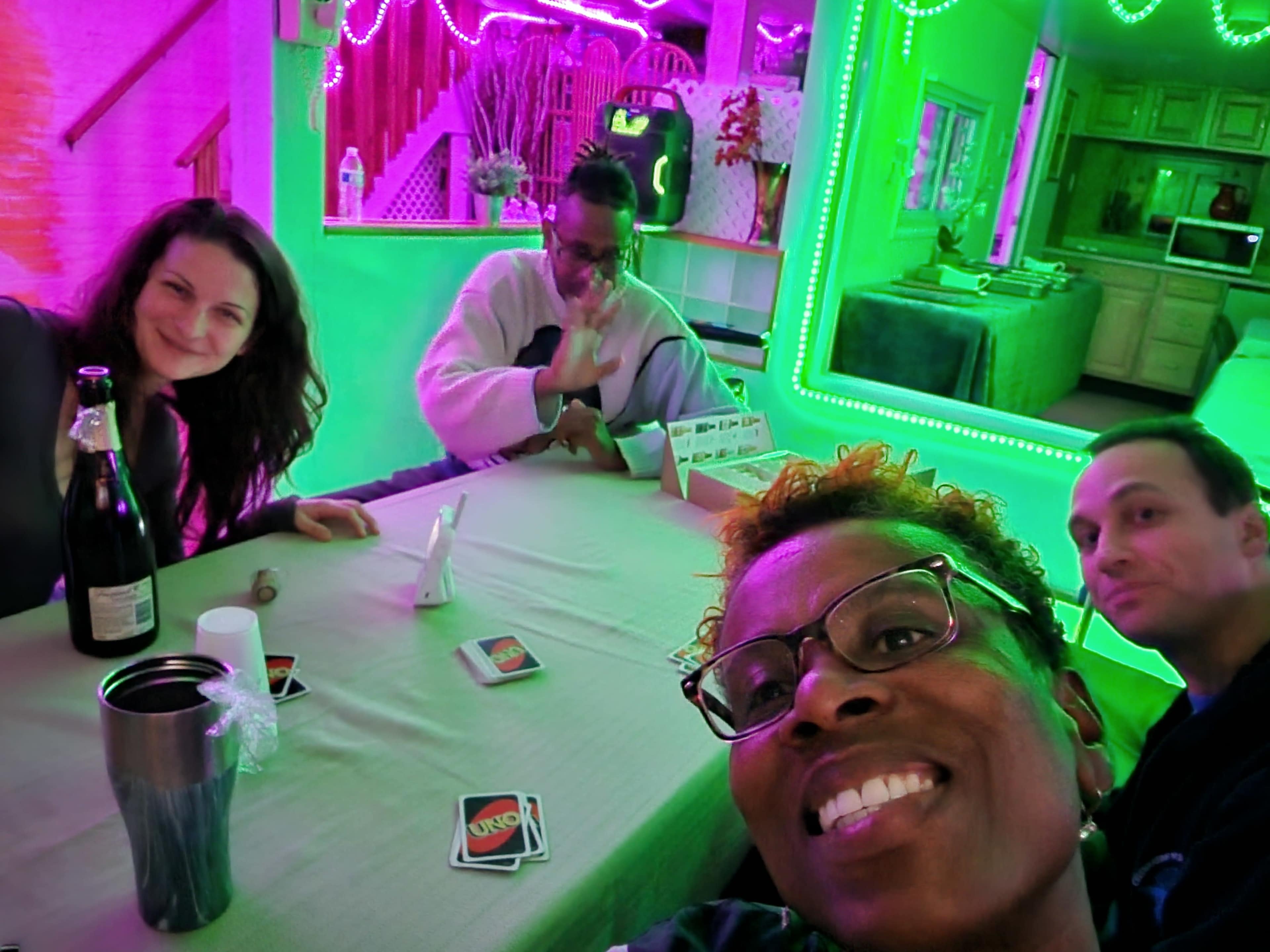 A group of four people is gathered around a table, with vibrant green lights illuminating the room, and an Uno game set up in front of them.