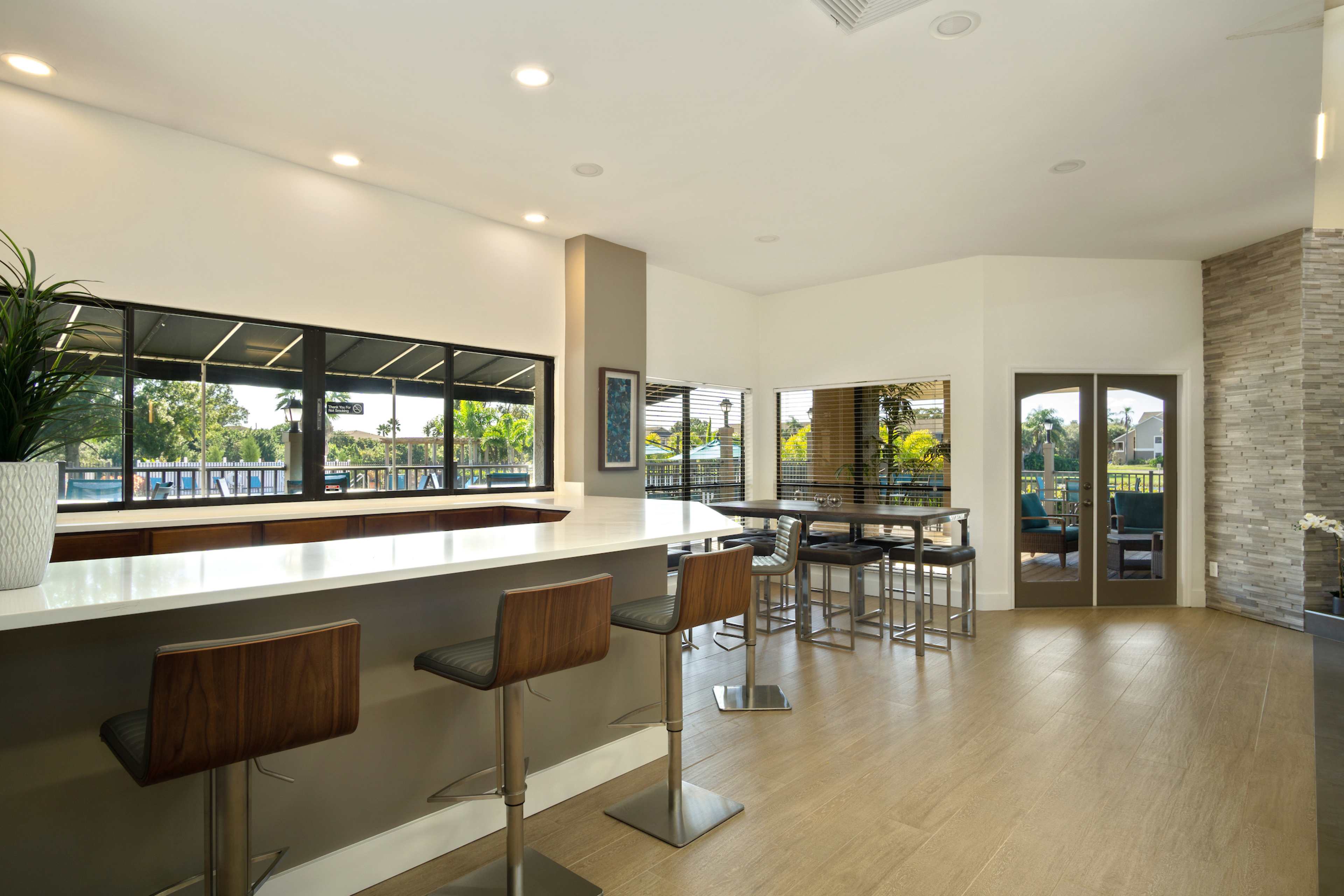 The image depicts a modern interior space with a long countertop, bar stools, large windows overlooking greenery, and a seating area with additional tables and chairs.