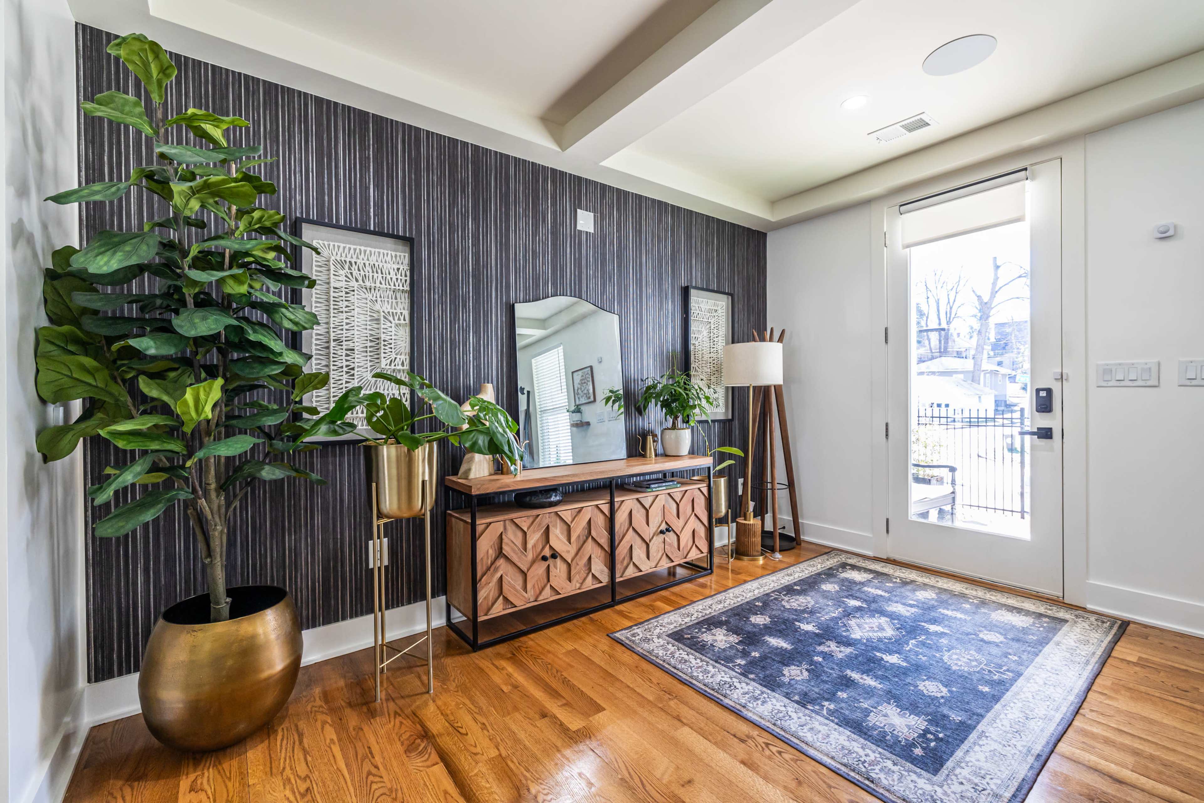 The image shows a modern entryway featuring a console table with decorative items, a large mirror, a plant in a pot, and a front door leading to an outdoor area.