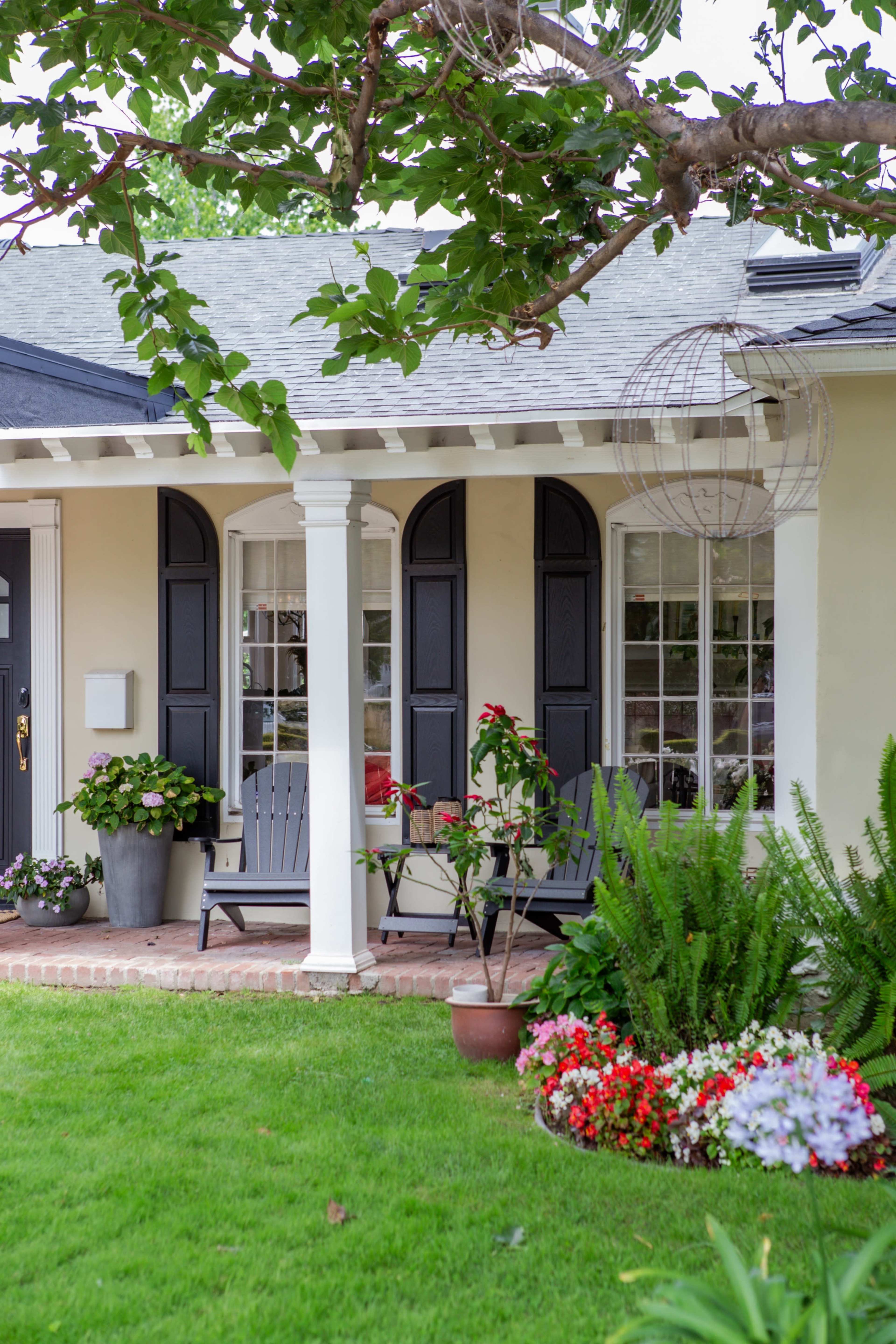 The image shows a front porch of a house with black shutters, two chairs, a variety of potted plants, and a grassy lawn.