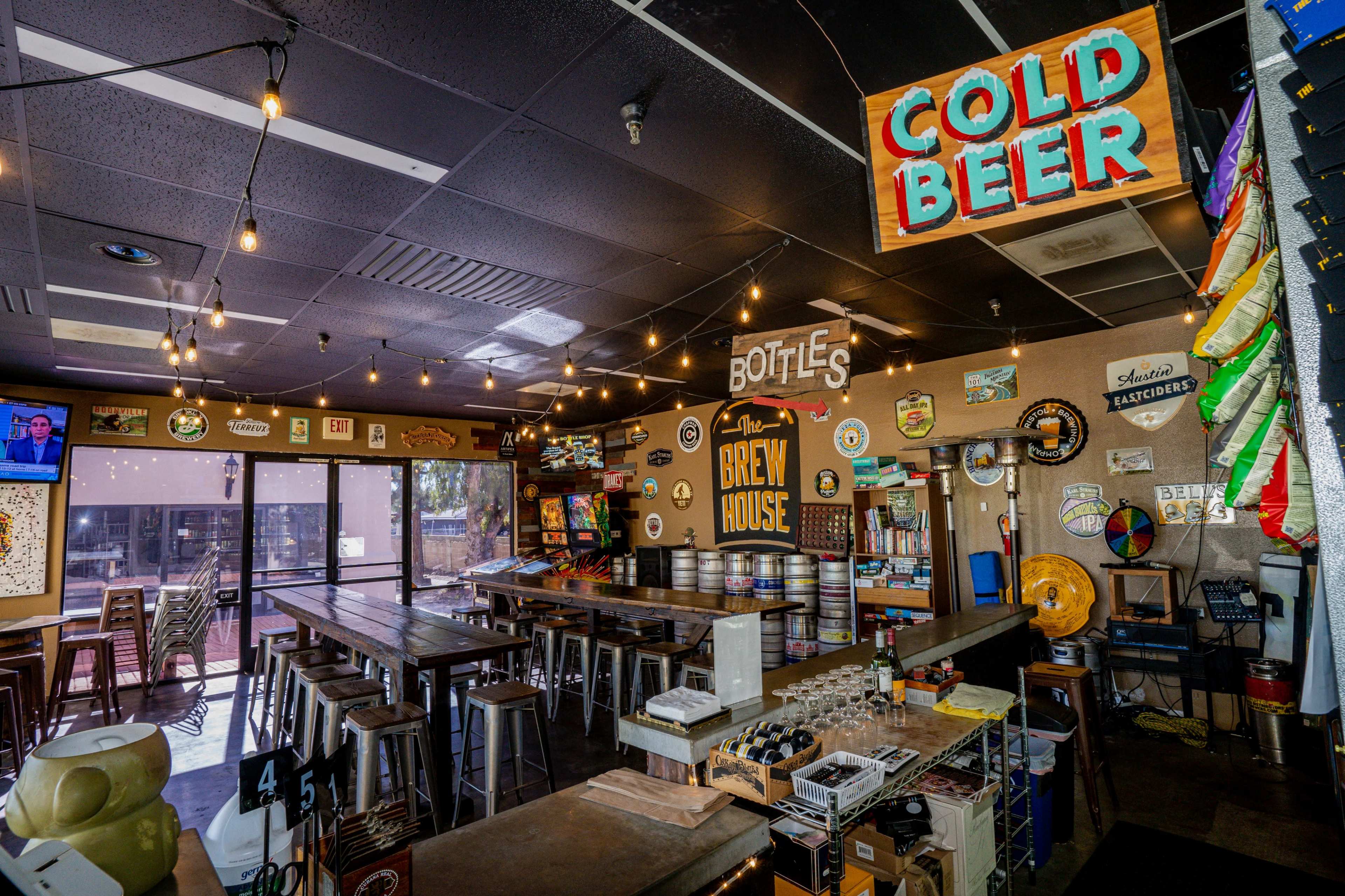 A bar interior with a long wooden table, kegs stacked on the walls, and colorful signs reading "COLD BEER" and "BOTTLES."