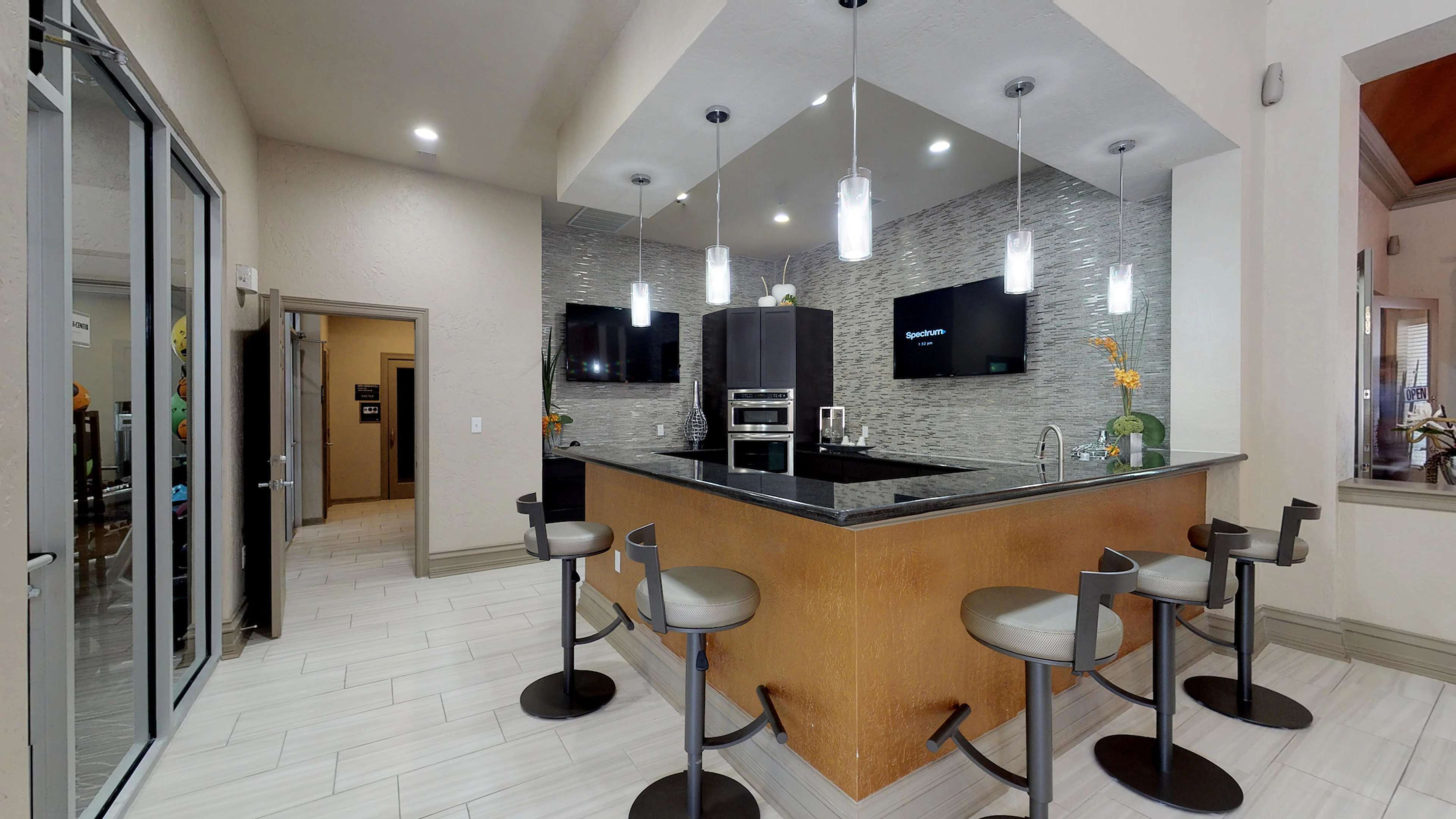 A modern kitchen with a bar counter, four stools, and two wall-mounted televisions.