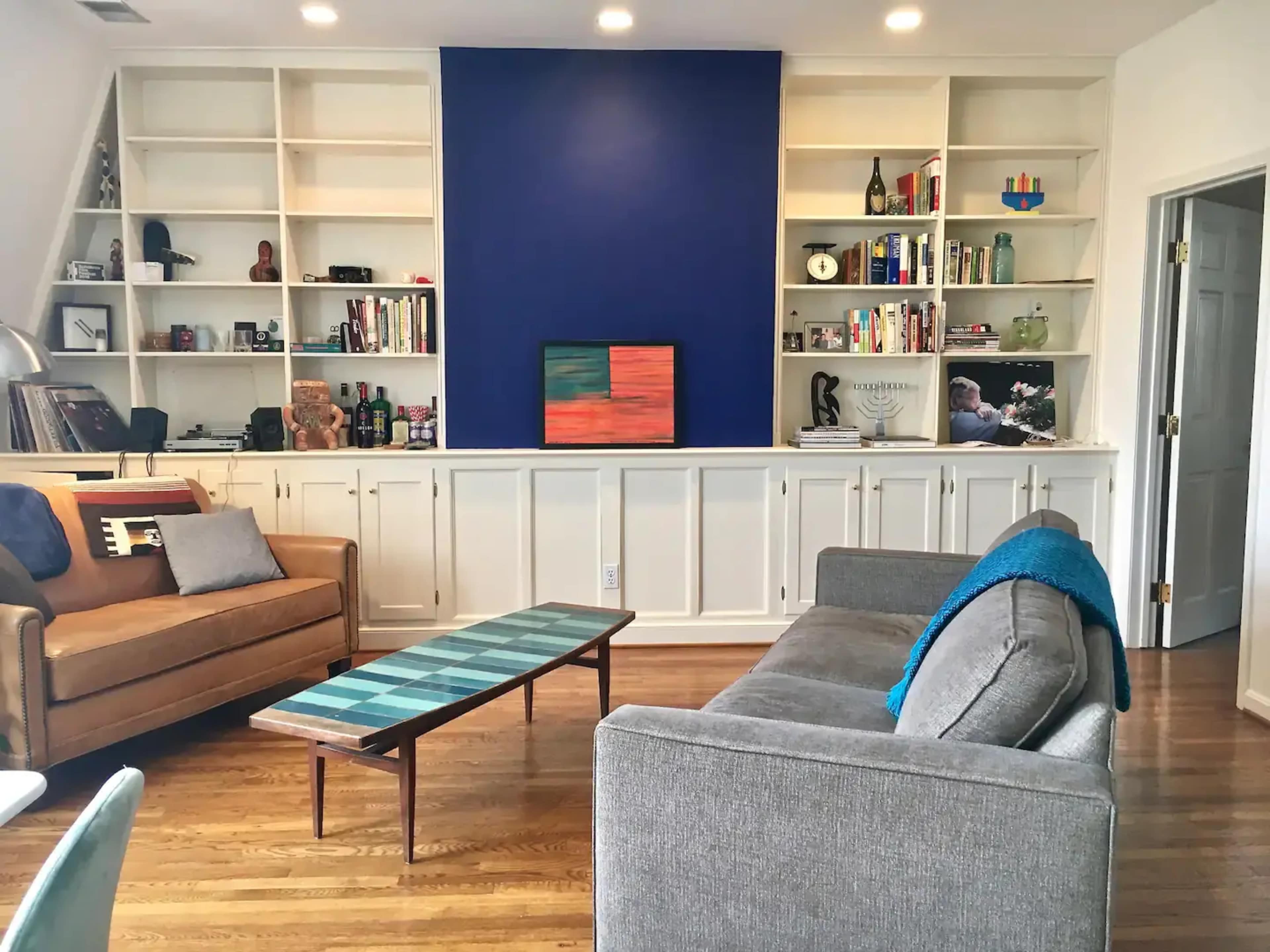A living room with two sofas facing a low coffee table, a bookshelf filled with various items, and a blue accent wall.
