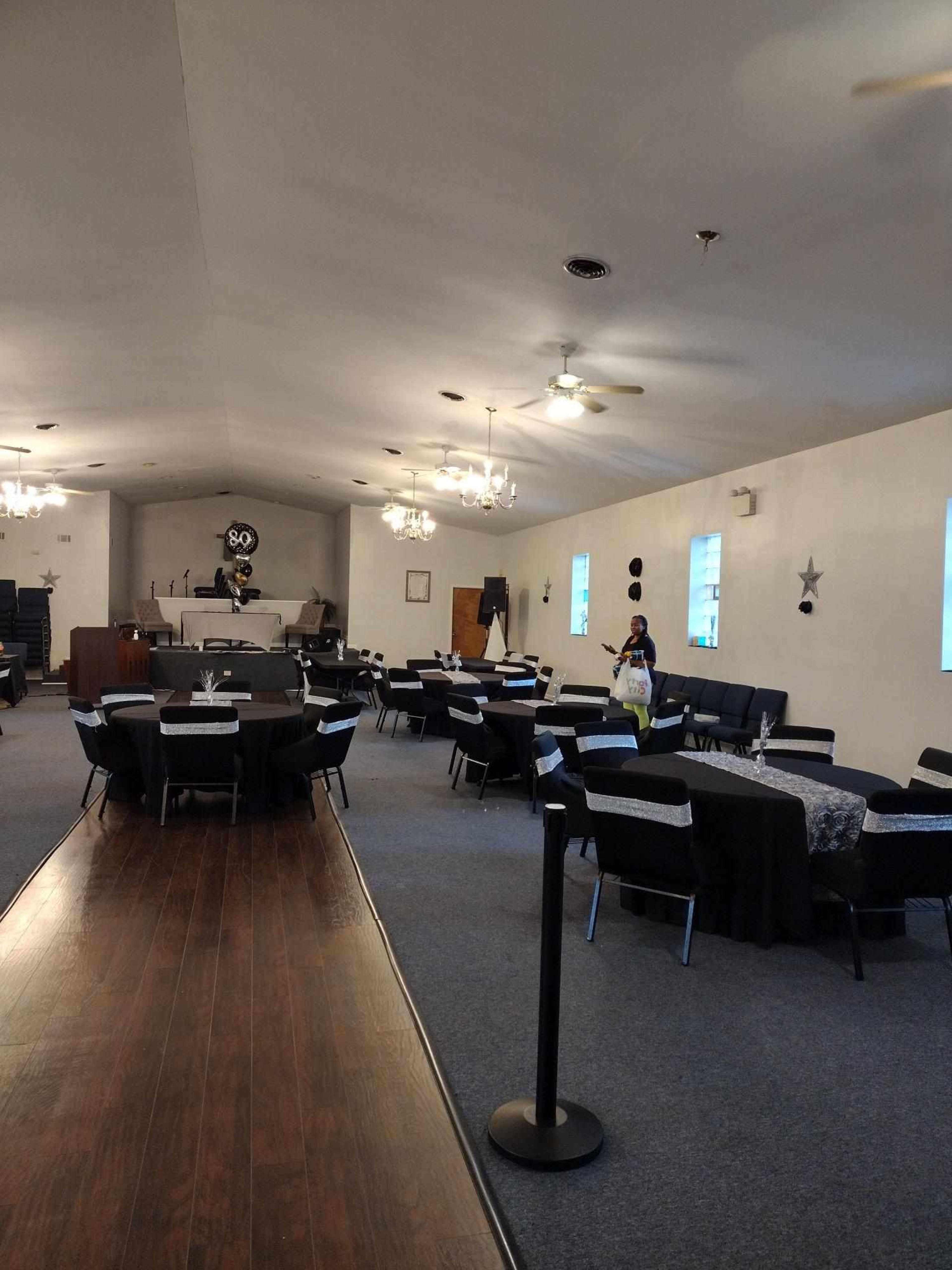 A spacious event hall set up with round tables covered in black cloth and decorated for a gathering, with a stage area at the back.