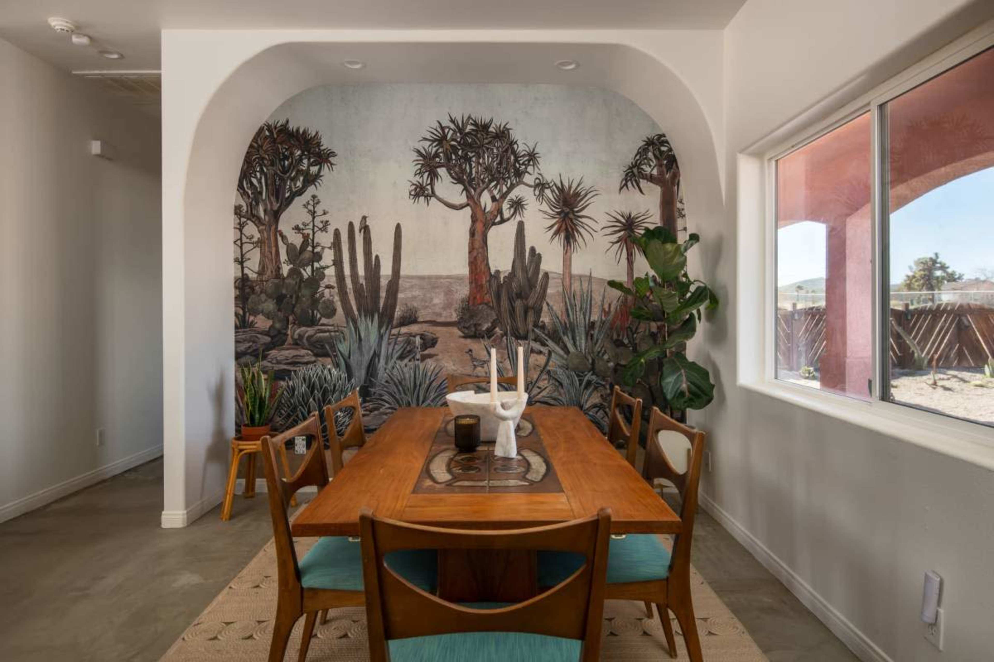 A dining area with a wooden table surrounded by chairs, featuring a mural of desert vegetation on the wall and a large window to the side.