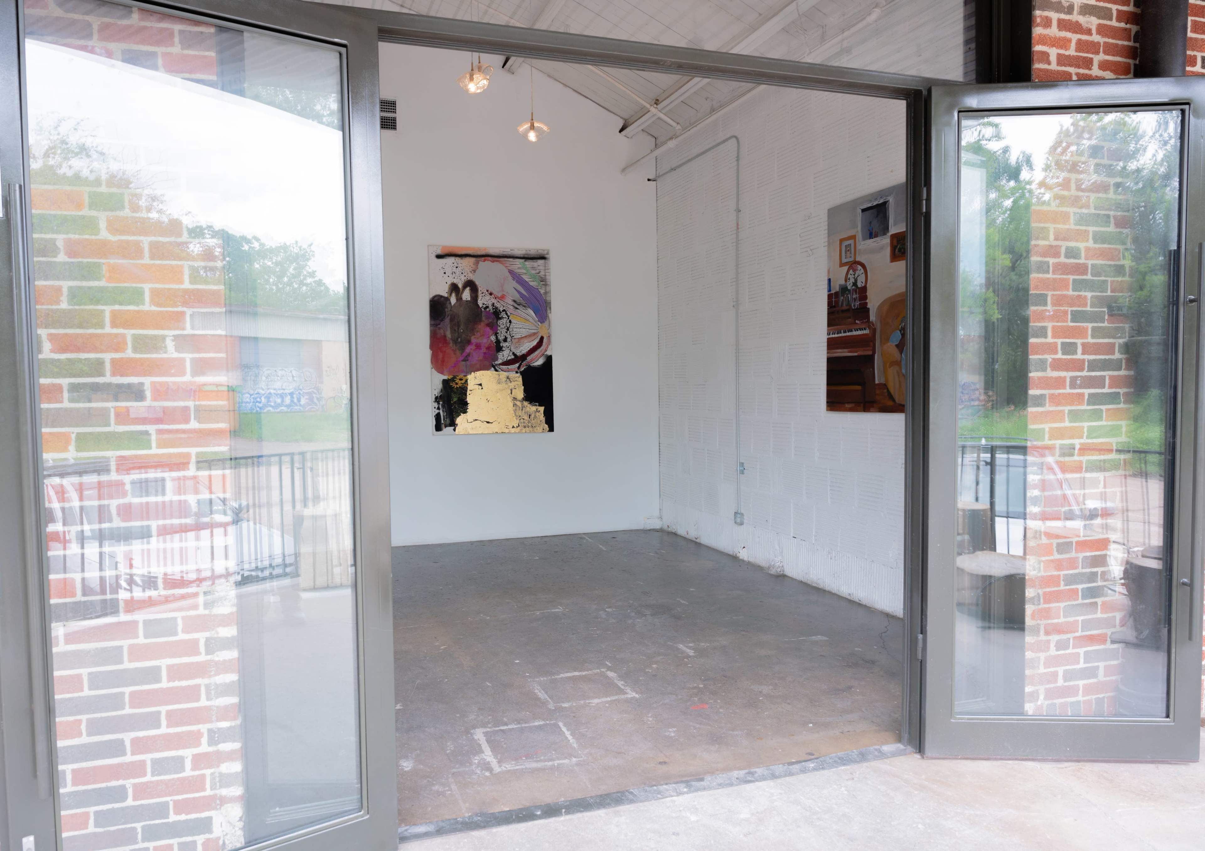 The image shows an empty art gallery space with open double doors, revealing a bare concrete floor and art pieces hanging on the white walls.