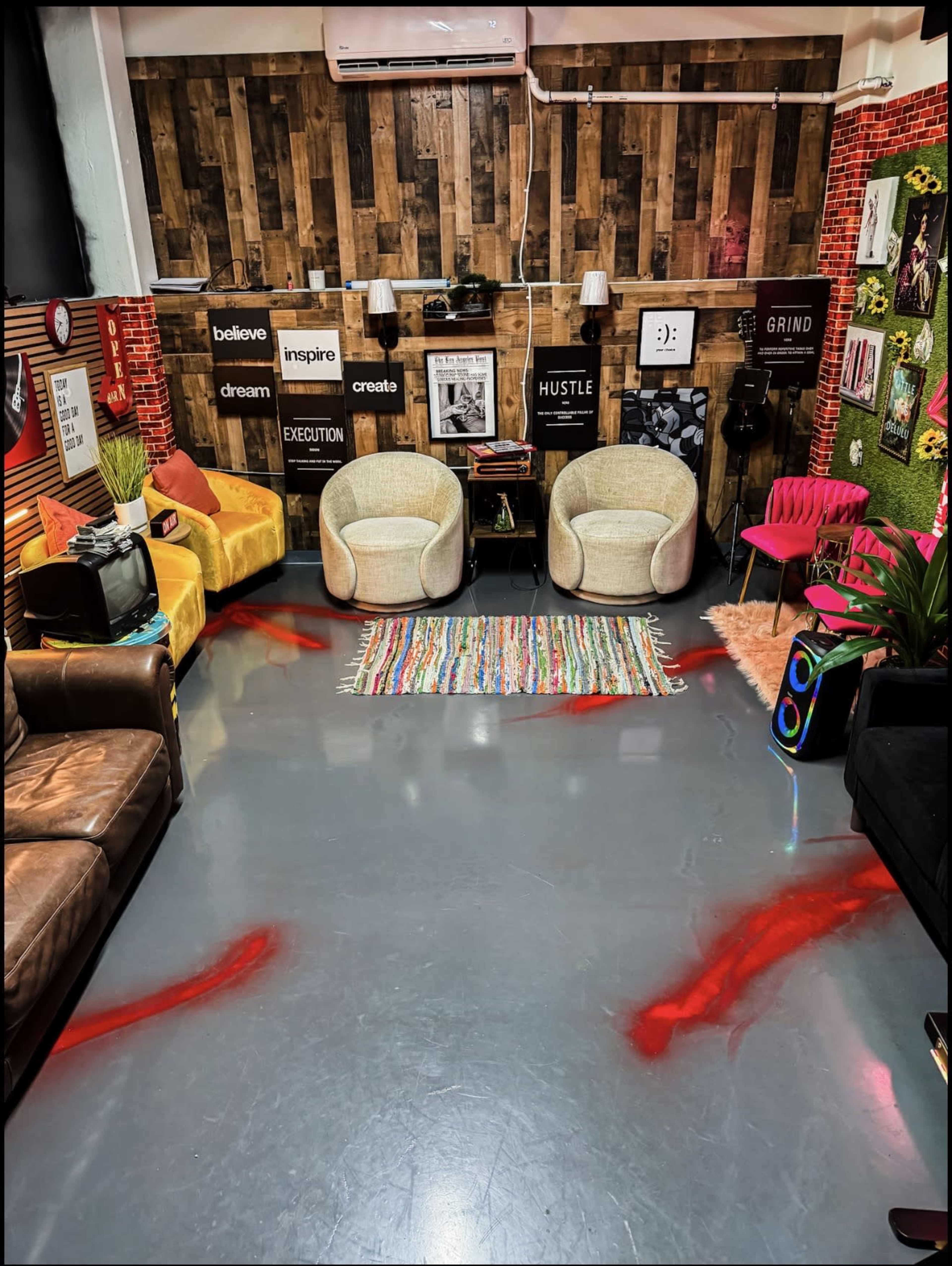 The image shows a stylish, modern coworking space with colorful furniture, including two armchairs and a pink chair, surrounded by decor featuring motivational posters and plants.