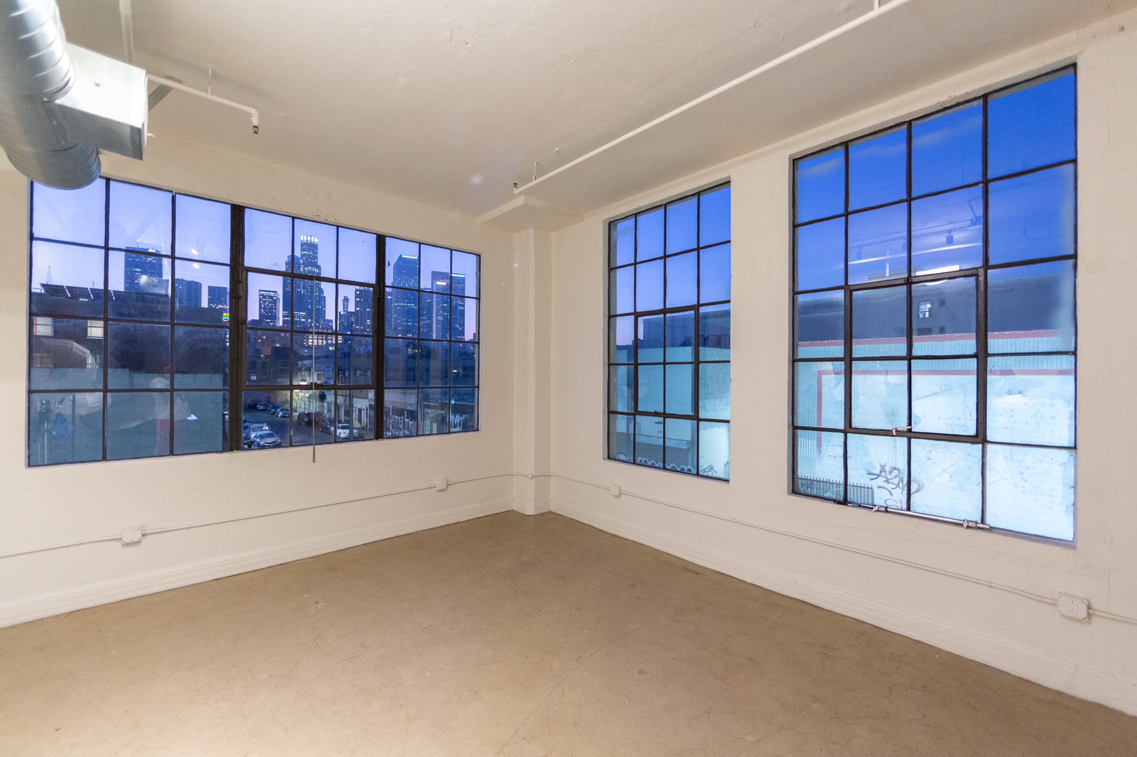 A spacious room with large windows overlooking a city skyline at dusk.