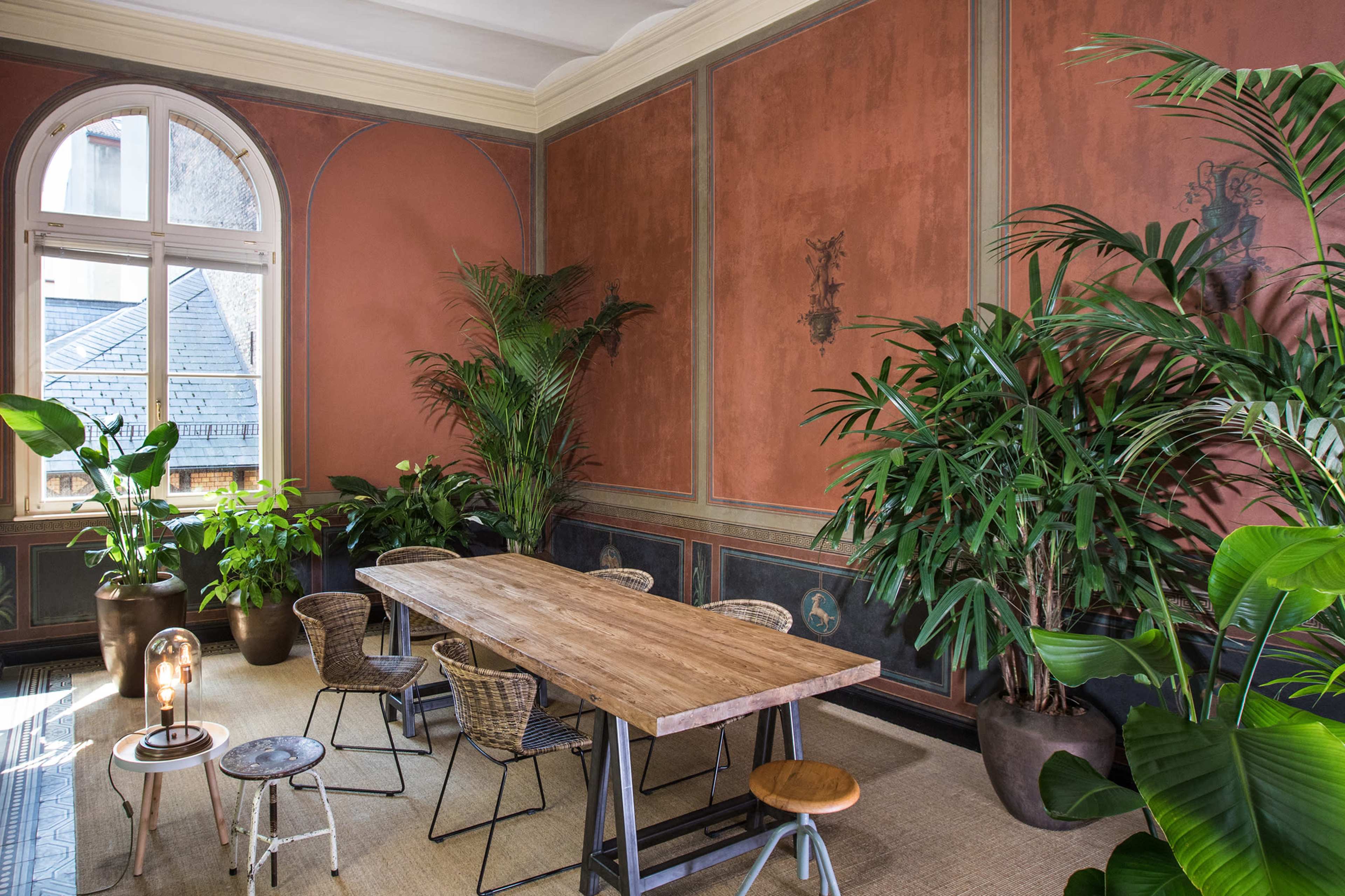 The image shows a spacious room with a wooden dining table surrounded by various plants, featuring decorative walls and large windows allowing natural light.