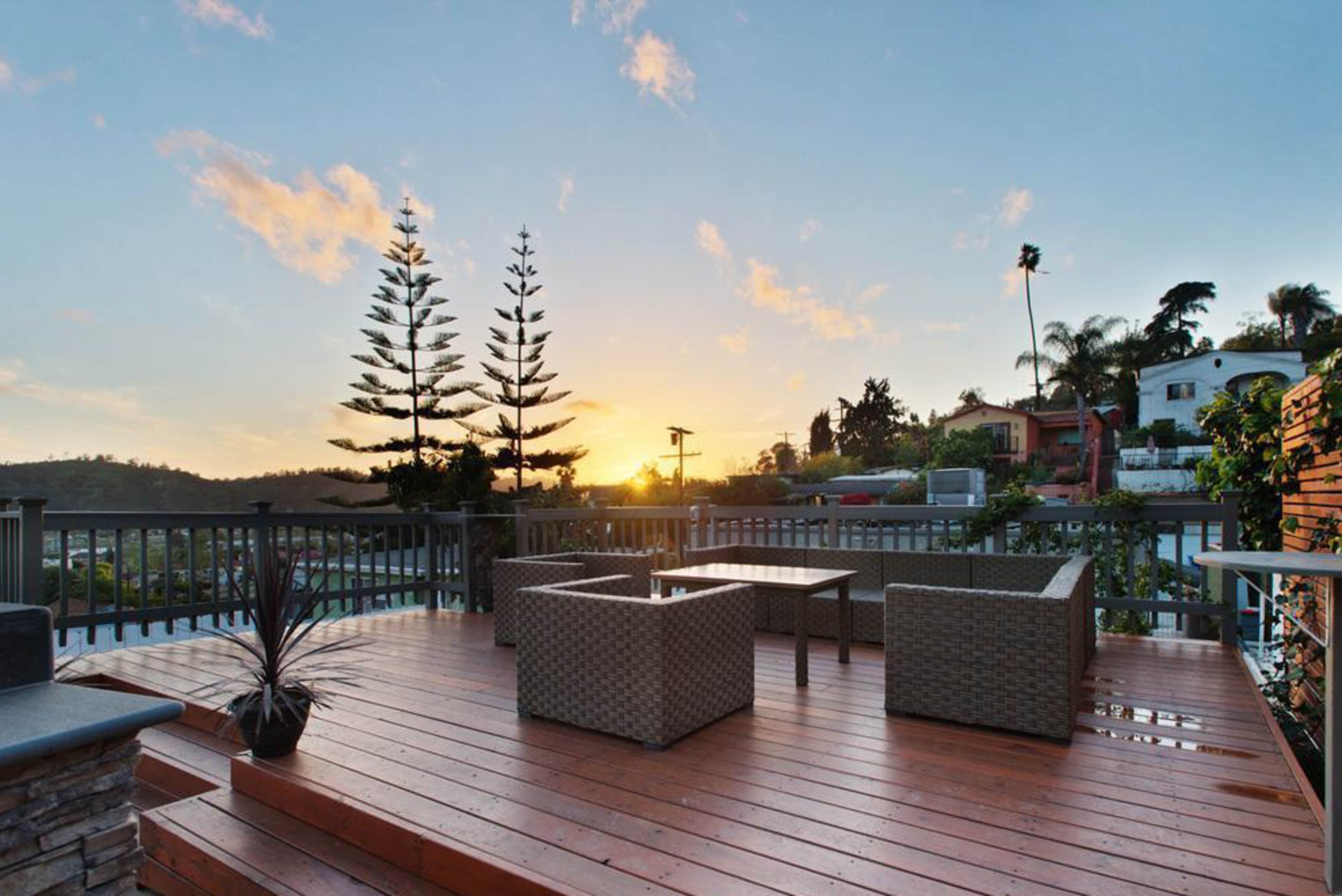 A wooden deck features two chairs and a table, set against a backdrop of a sunset with palm trees and houses in the distance.