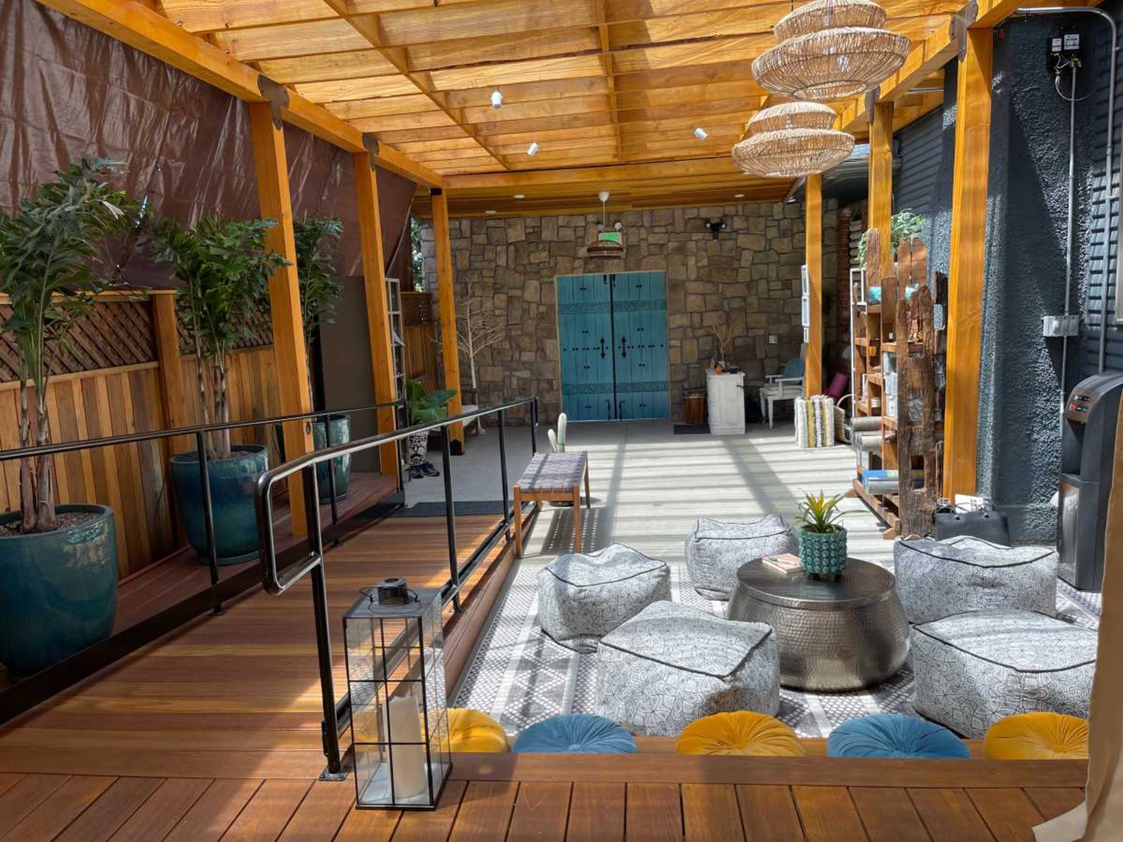 The image shows a spacious, modern indoor area with wooden beams, decorative plants, and various seating options arranged around a central table.