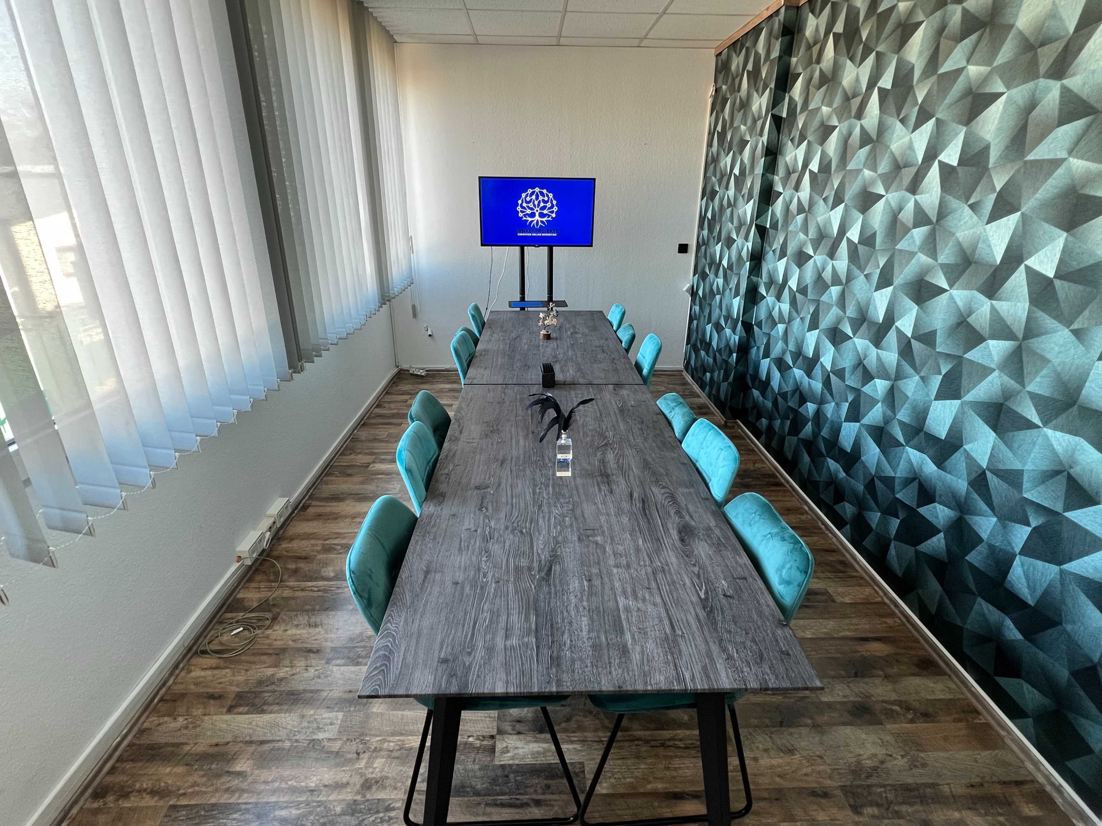 The image shows a meeting room with a long wooden table surrounded by teal chairs, and a television mounted on the wall displaying a logo.