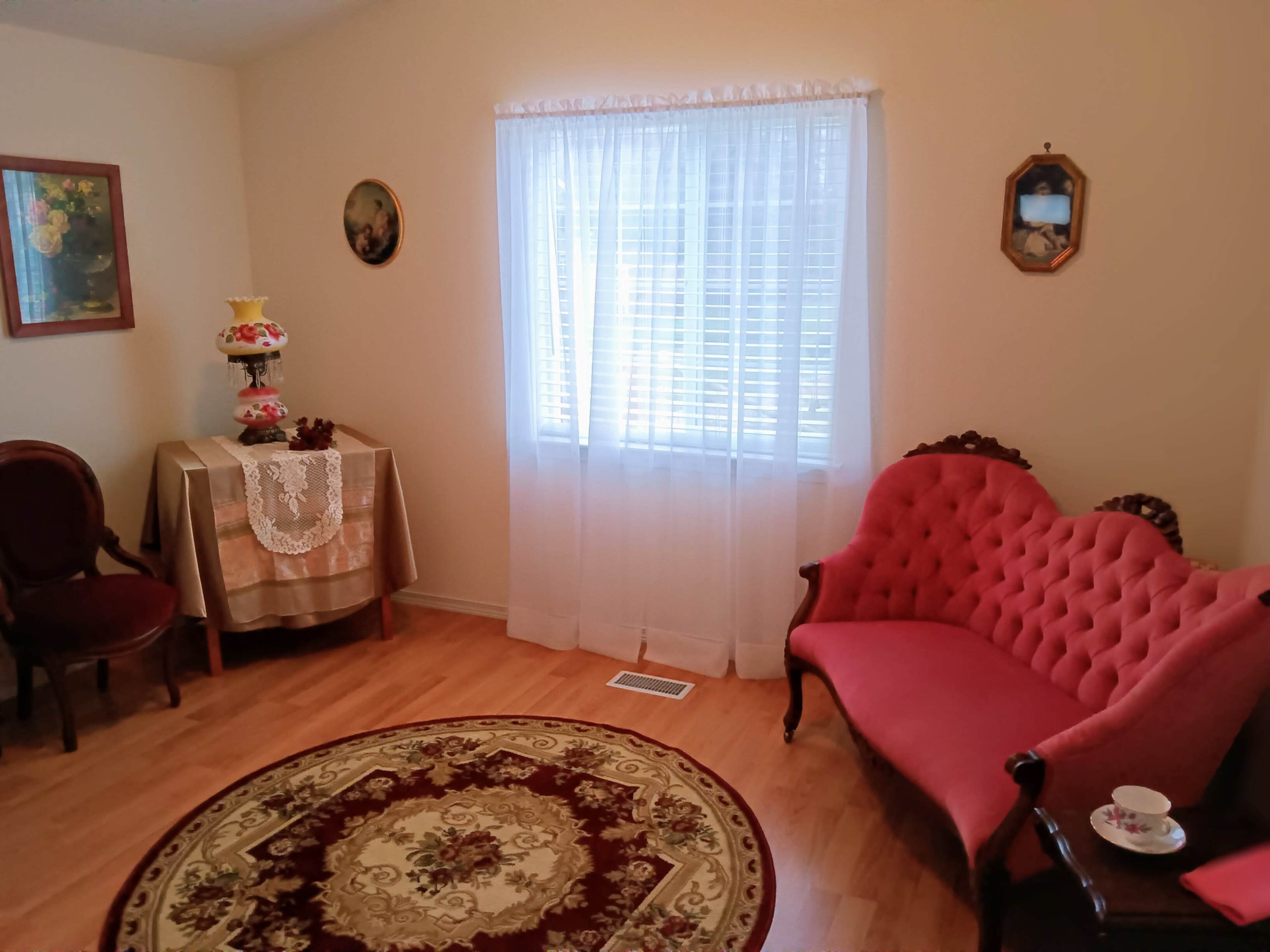 The room features a red tufted sofa, a round floral area rug, a table set with a lace tablecloth, and a large window draped with sheer curtains.
