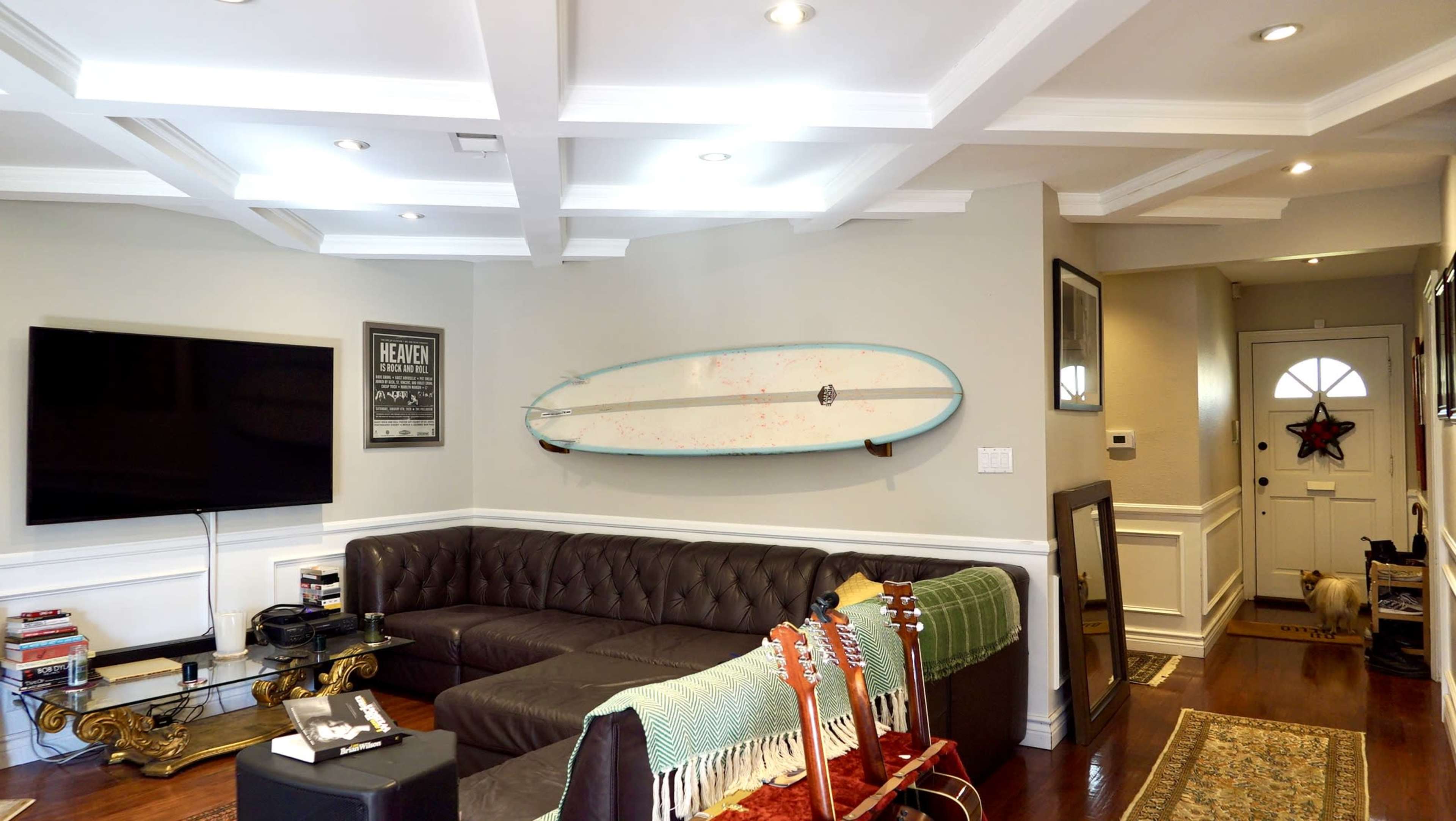 The image shows a modern living room featuring a brown leather couch, a surfboard mounted on the wall, a television, and a welcoming entrance with a door.