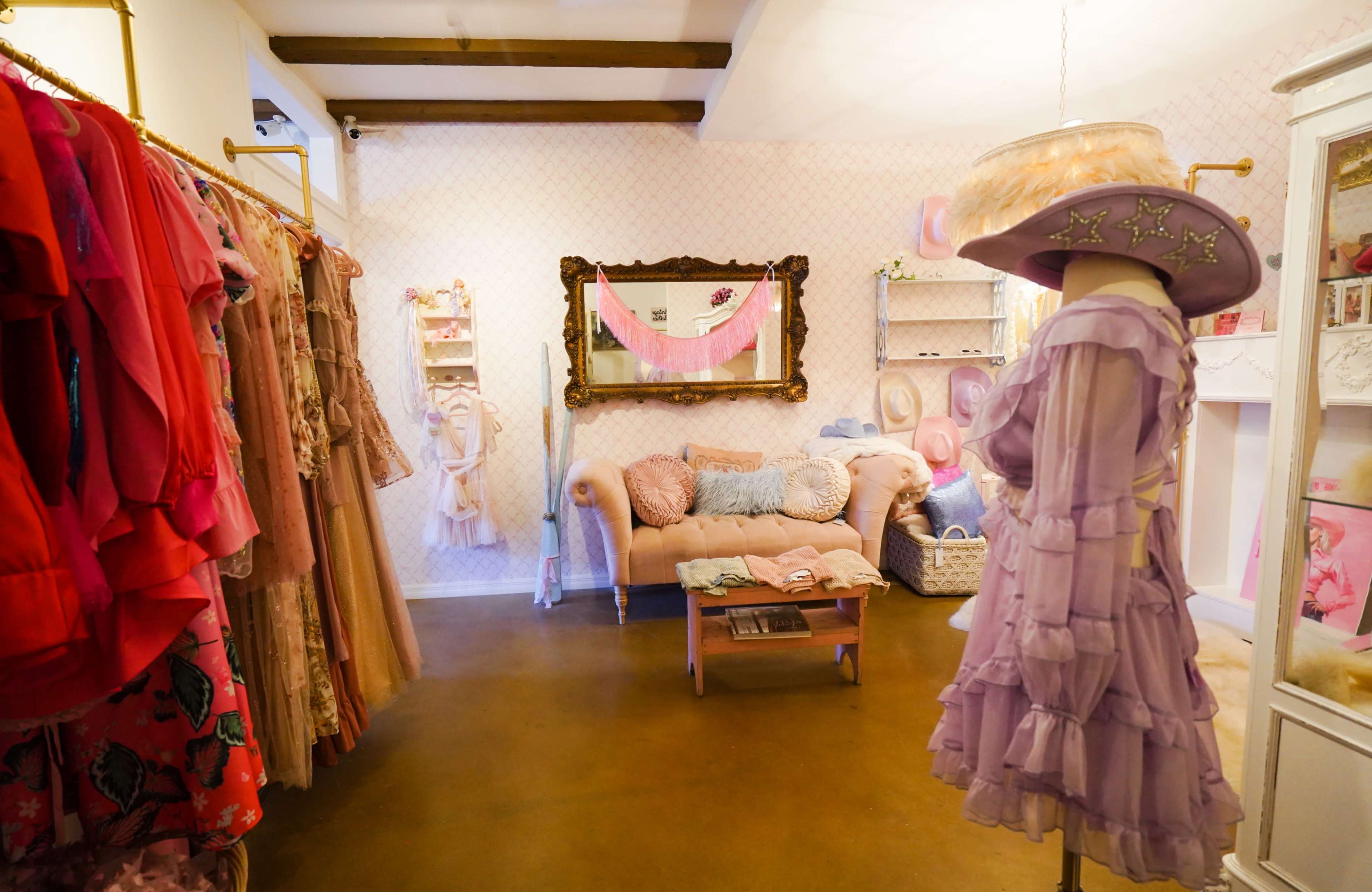 The image shows a boutique interior featuring a pastel color scheme, with racks of dresses, a vintage-style mirror, a pink sofa, and a mannequin displaying a lilac outfit.