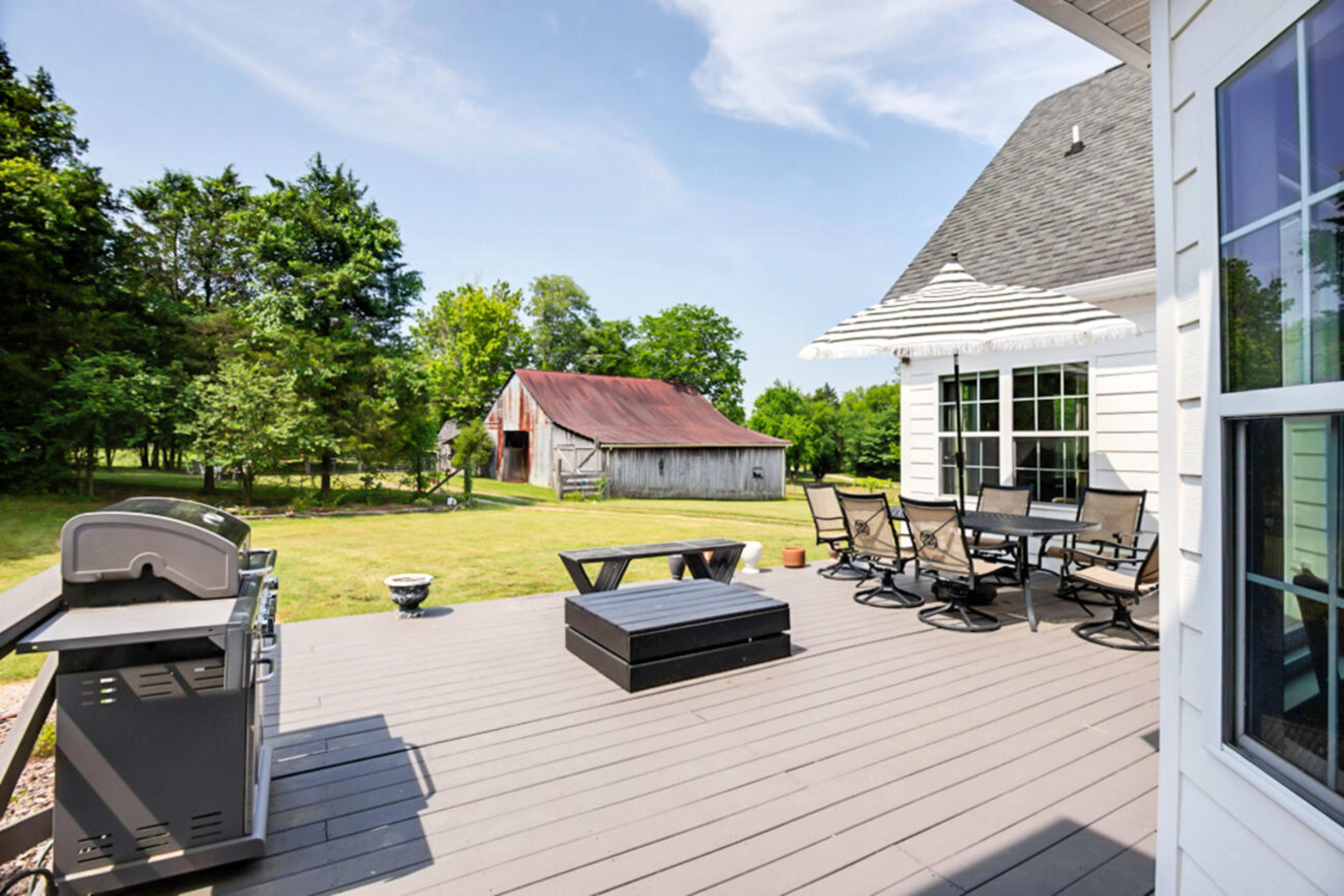 A spacious deck features a grill, patio furniture, and a view of a green lawn with a barn in the background.