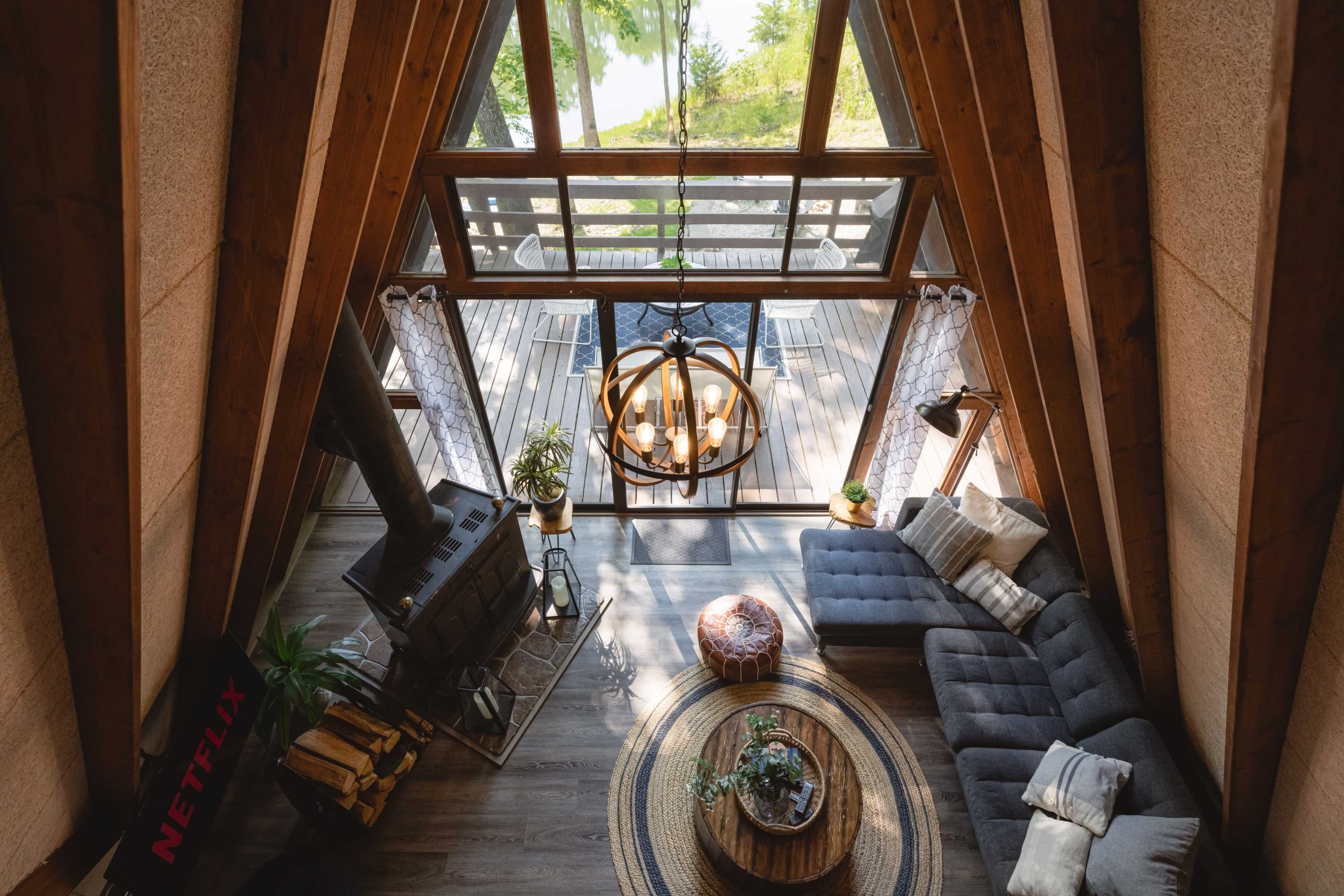 The image shows a modern A-frame cabin living area featuring a large sectional sofa, a circular coffee table with plants, a cozy wood stove, and large windows that provide a view of an outdoor deck.