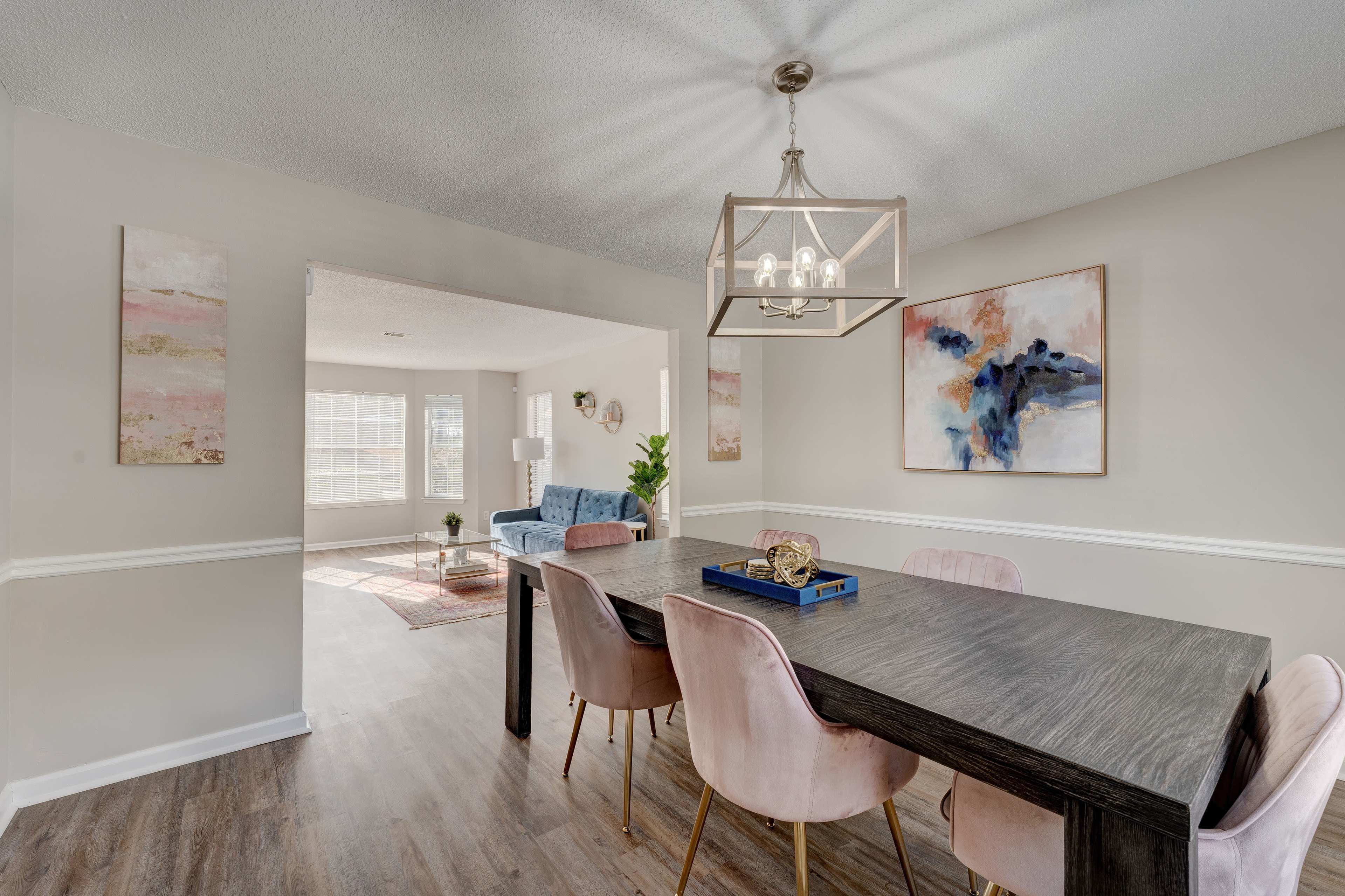 A modern dining area features a dark rectangular table with pink upholstered chairs and framed artwork on the walls, leading to a bright living space.