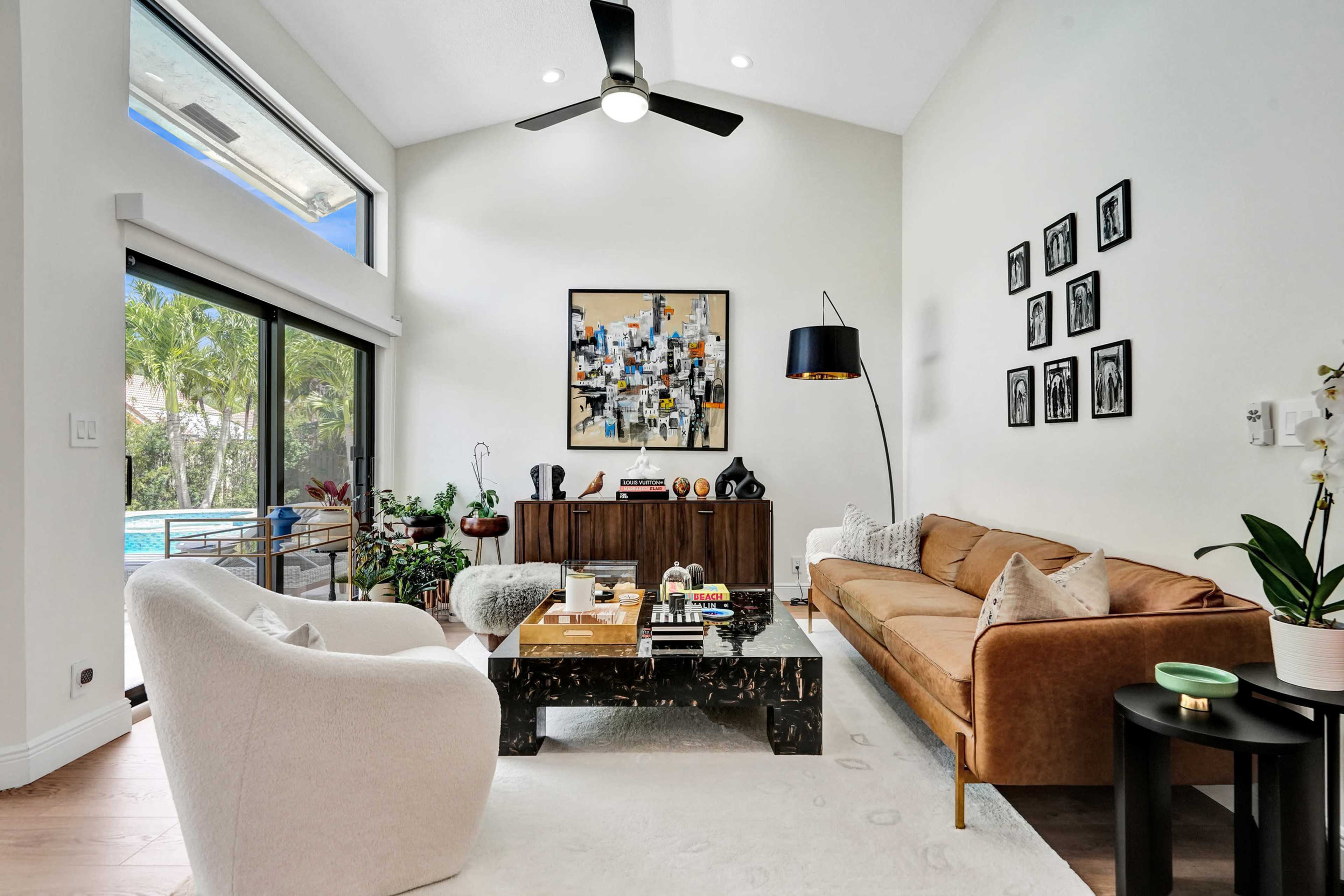 A modern living room features a brown sofa, a black coffee table, and a wall adorned with framed photos, alongside large windows that open to a view of greenery and a pool.